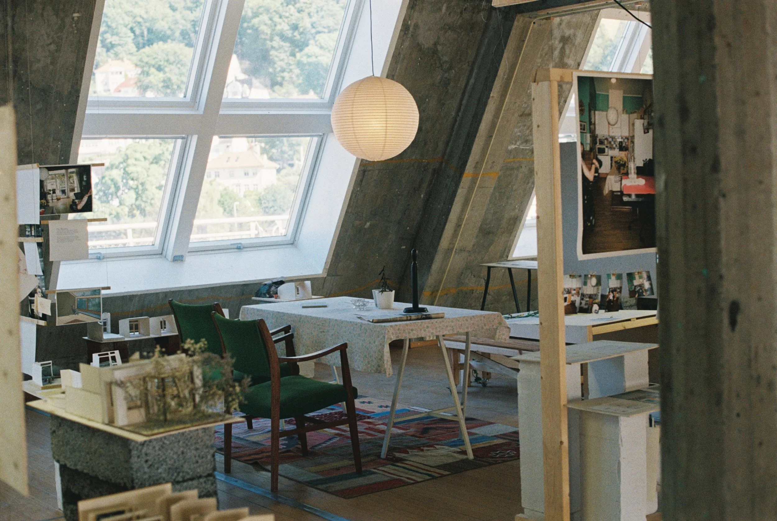 et åpent rom med skrå tak og store vinduer. et kjøkkenbort i midten med stoler rundt. en lampe henger fra taketog en fargerik matte under. fotografier og modeller kan skimtes