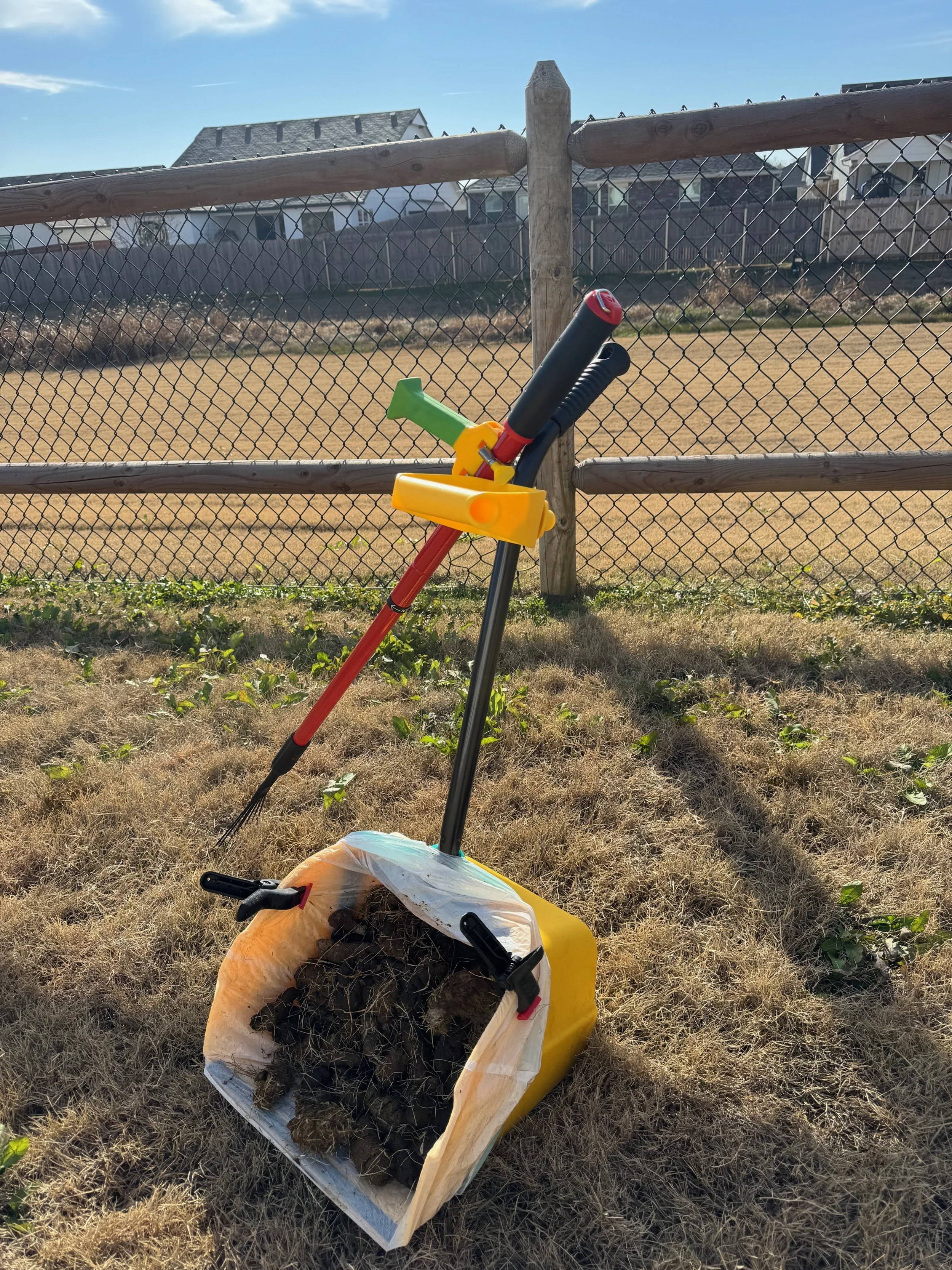 A small yellow trash bag filled with horse manure, a green toy rake, and a red and black toy pitchfork leaning against a wooden fence in a backyard.