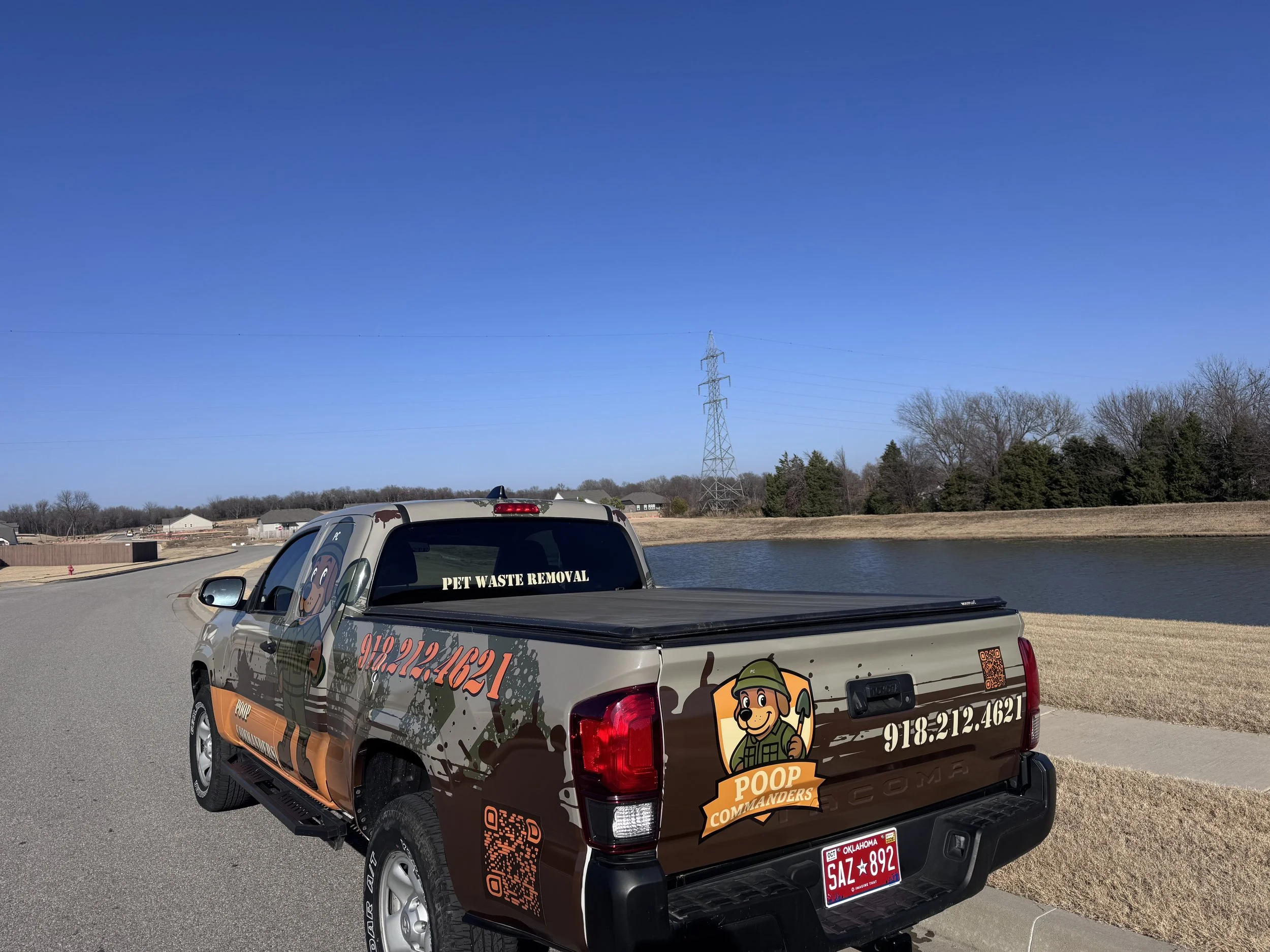 A pet waste removal company truck parked on a street near a pond with a clear blue sky in the background. The truck features graphics including a cartoon dog dressed as a soldier, phone number, QR code, and company logo.