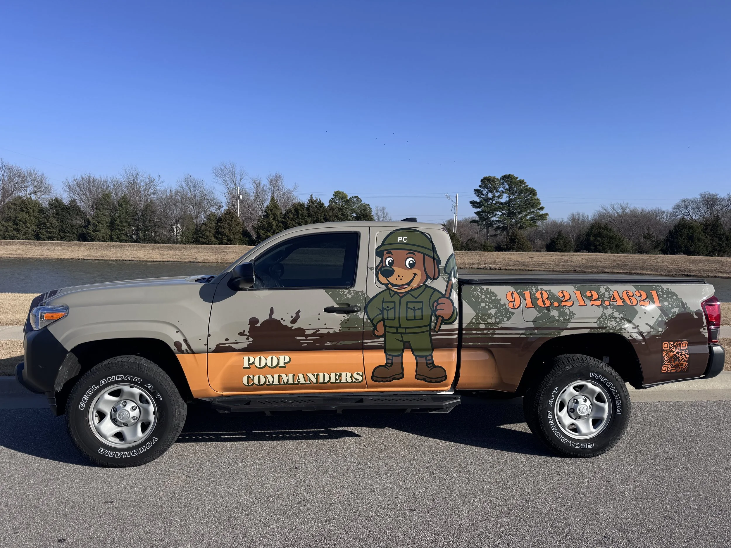 A pickup truck with a logo of a cartoon dog dressed as a military soldier, holding a shovel. The truck advertises 'Poop Commanders' with a phone number and QR code on the side.