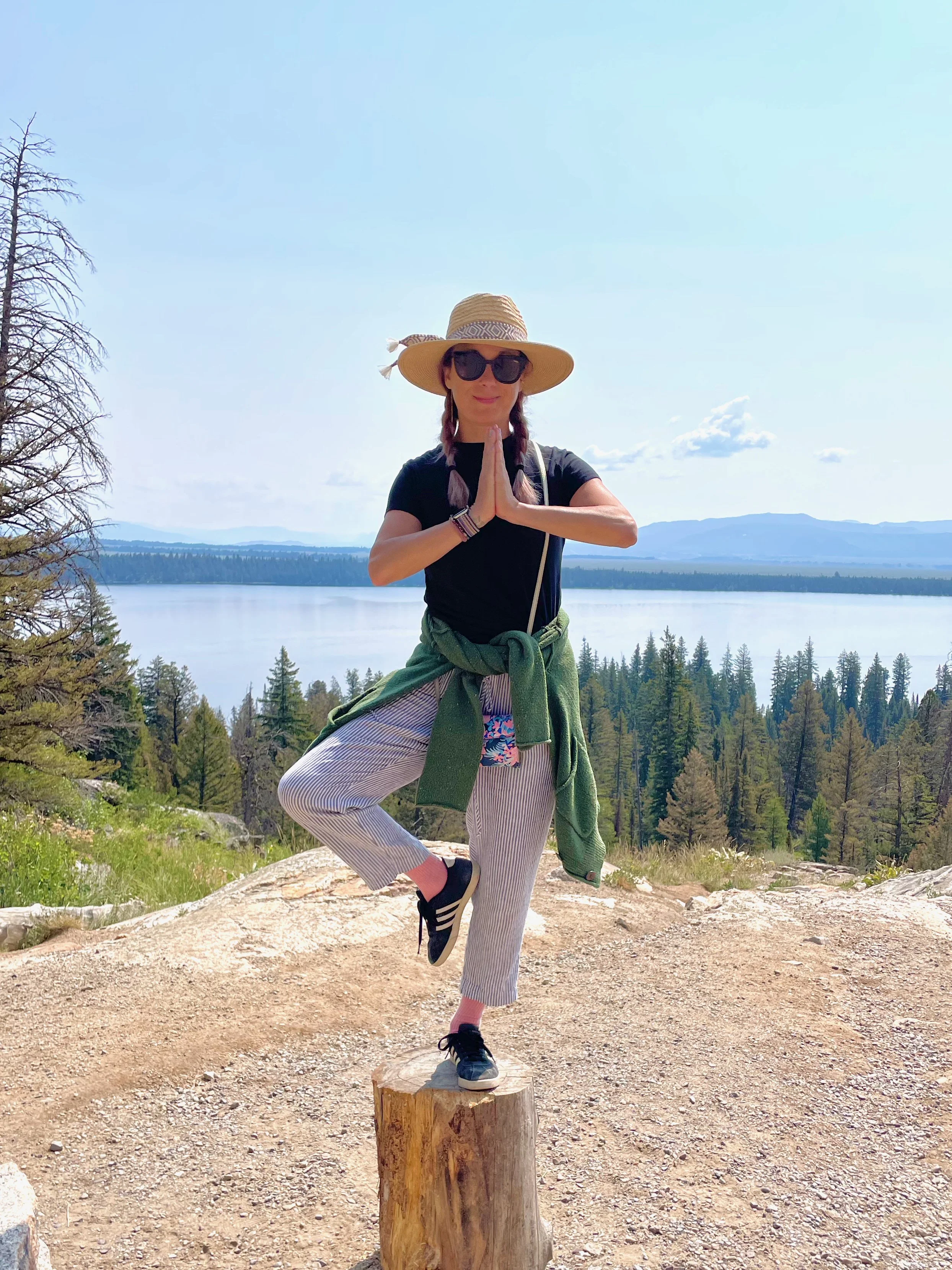 Roberta practicing yoga outdoors on a tree stump near a lake, wearing a wide-brimmed hat, sunglasses, black sneakers, with a green jacket tied around her waist, hands in a prayer position. Yellowstone National Park, Jenny Lake