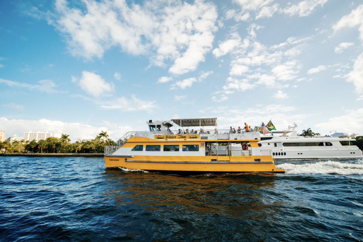 A yellow boat named 'Water Taxi' on the water with a white yacht in the background, under a partly cloudy sky, with trees and buildings along the shoreline.