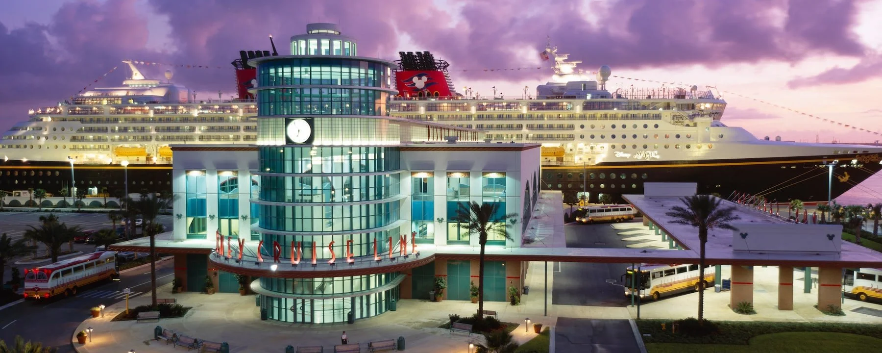 The Disney Cruise Line ship docked at the port with the city skyline and purple sunset sky in the background. The cruise terminal building has a large glass front with a clock and the words "The Cruise Line" in red lights. Palm trees and buses are visible in the foreground.
