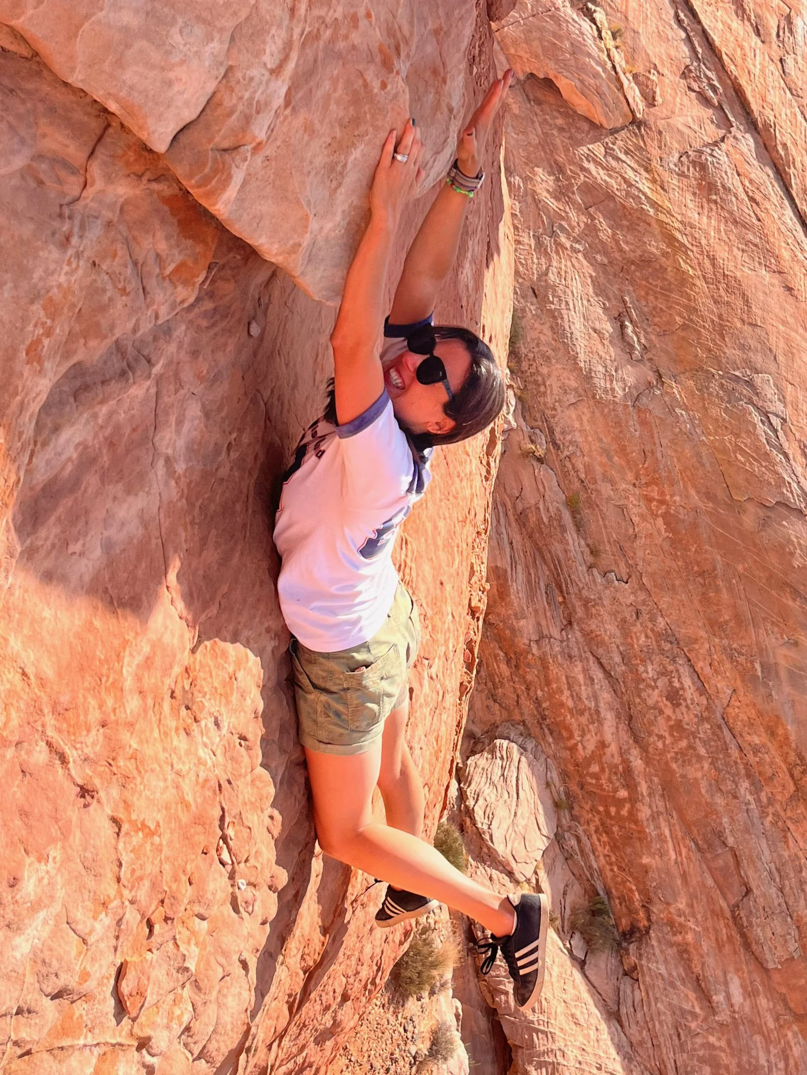 A woman rock climbing on a reddish-brown cliff with a joyous expression, wearing sunglasses, a white t-shirt, camouflage shorts, and sneakers.
