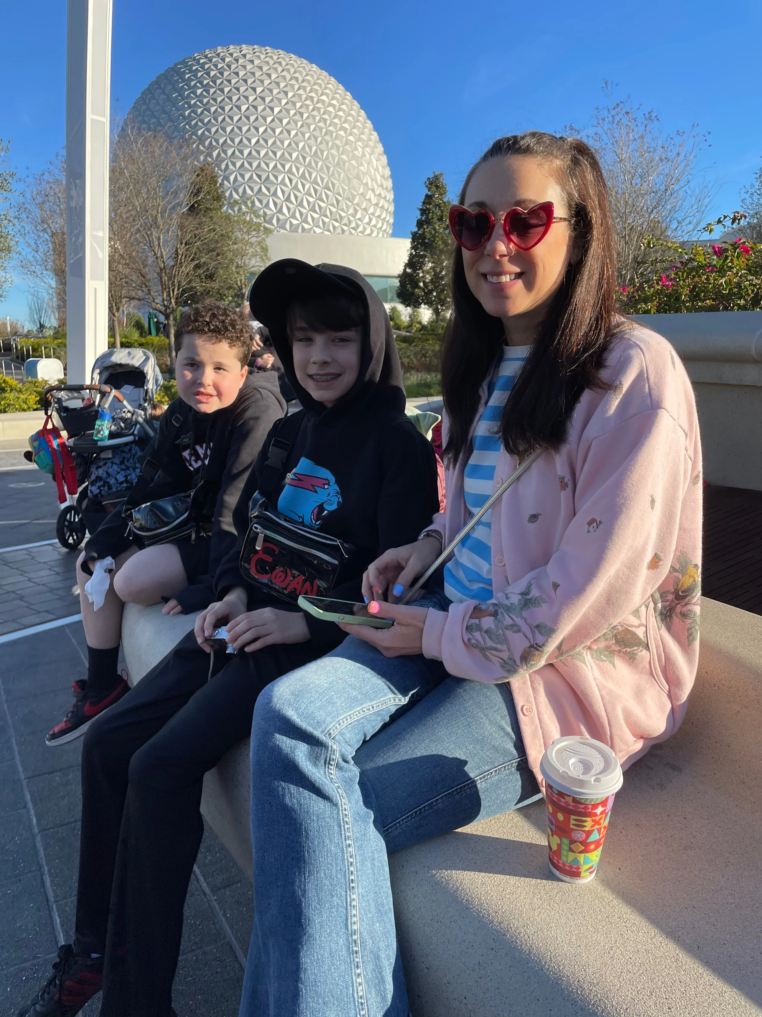 Roberta with heart-shaped sunglasses sitting on a bench with two young boys, one wearing a hoodie and the other a jacket, in front of Epcot's Spaceship Earth geodesic sphere at Disney World.