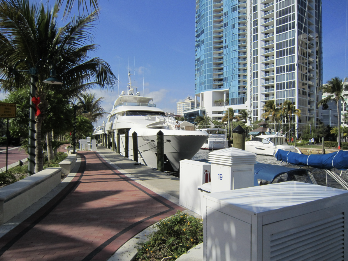 Marina with several yachts docked along a brick-paved walkway, tall modern buildings in the background, palm trees lining the pathway, clear blue sky.