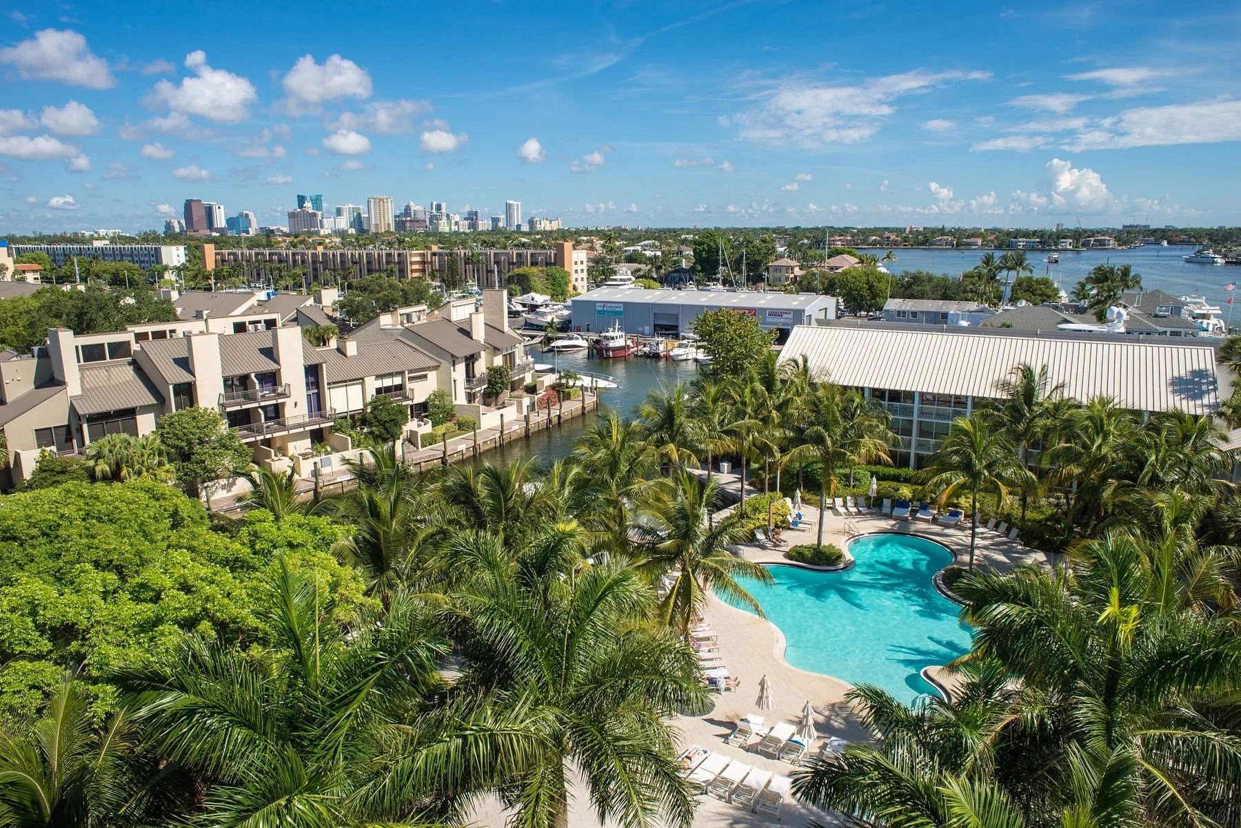 A tropical resort with a swimming pool surrounded by palm trees, and residential buildings with boats docked on a waterway, with a city skyline and blue sky in the background.