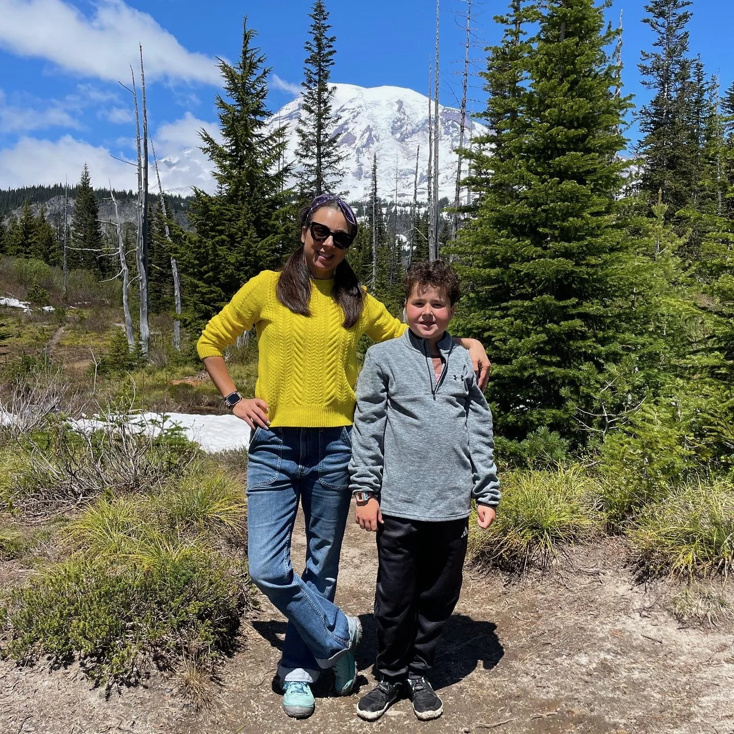 Roberta and a young boy standing outdoors on a trail in a forest with evergreen trees and snow patches, Mount Rainier snow-capped mountain in the background on a sunny day.