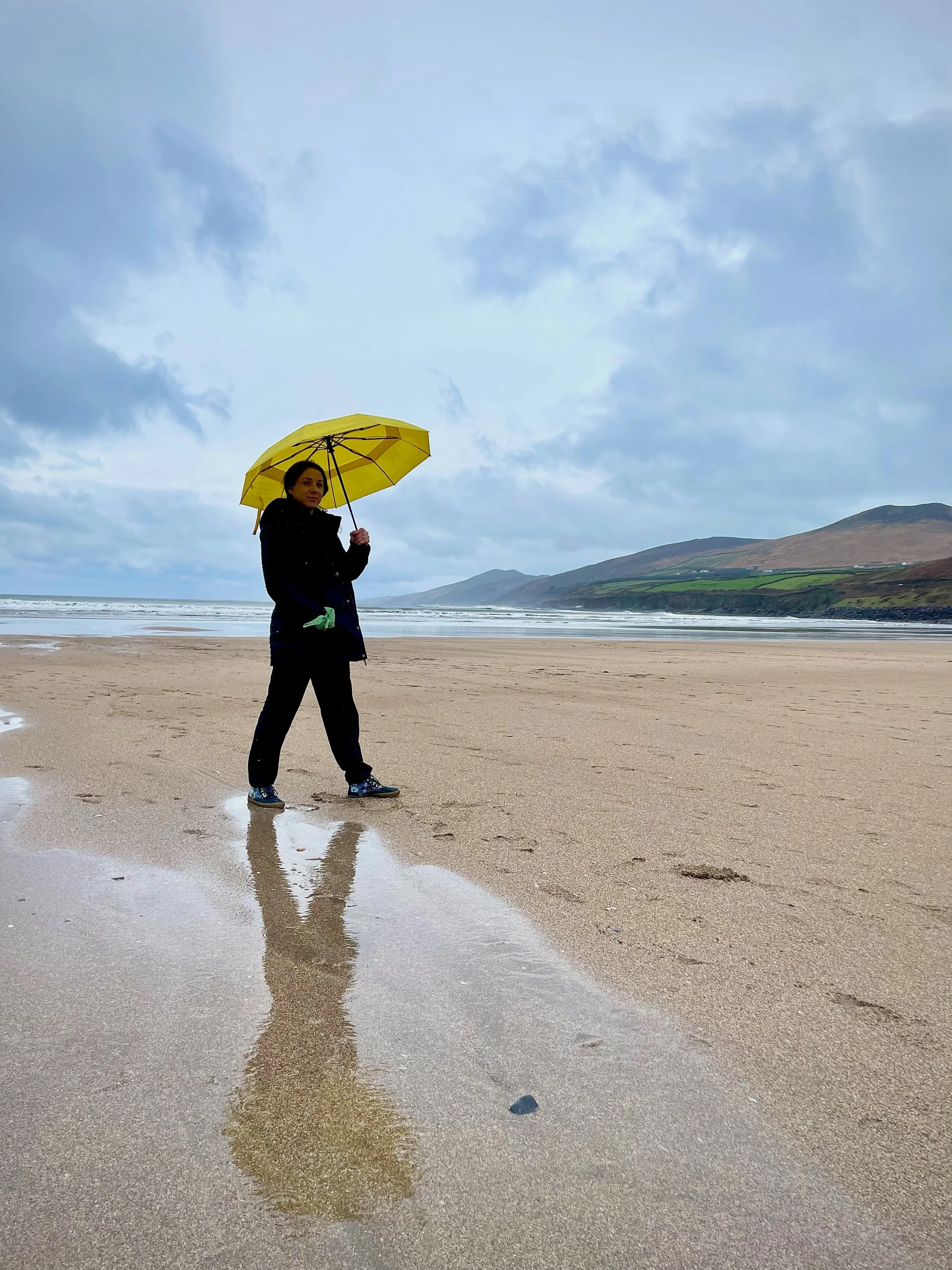 A woman holding a yellow umbrella walks on a rainy beach with wet sand and reflections, clouds overhead, and rolling hills in the distance.