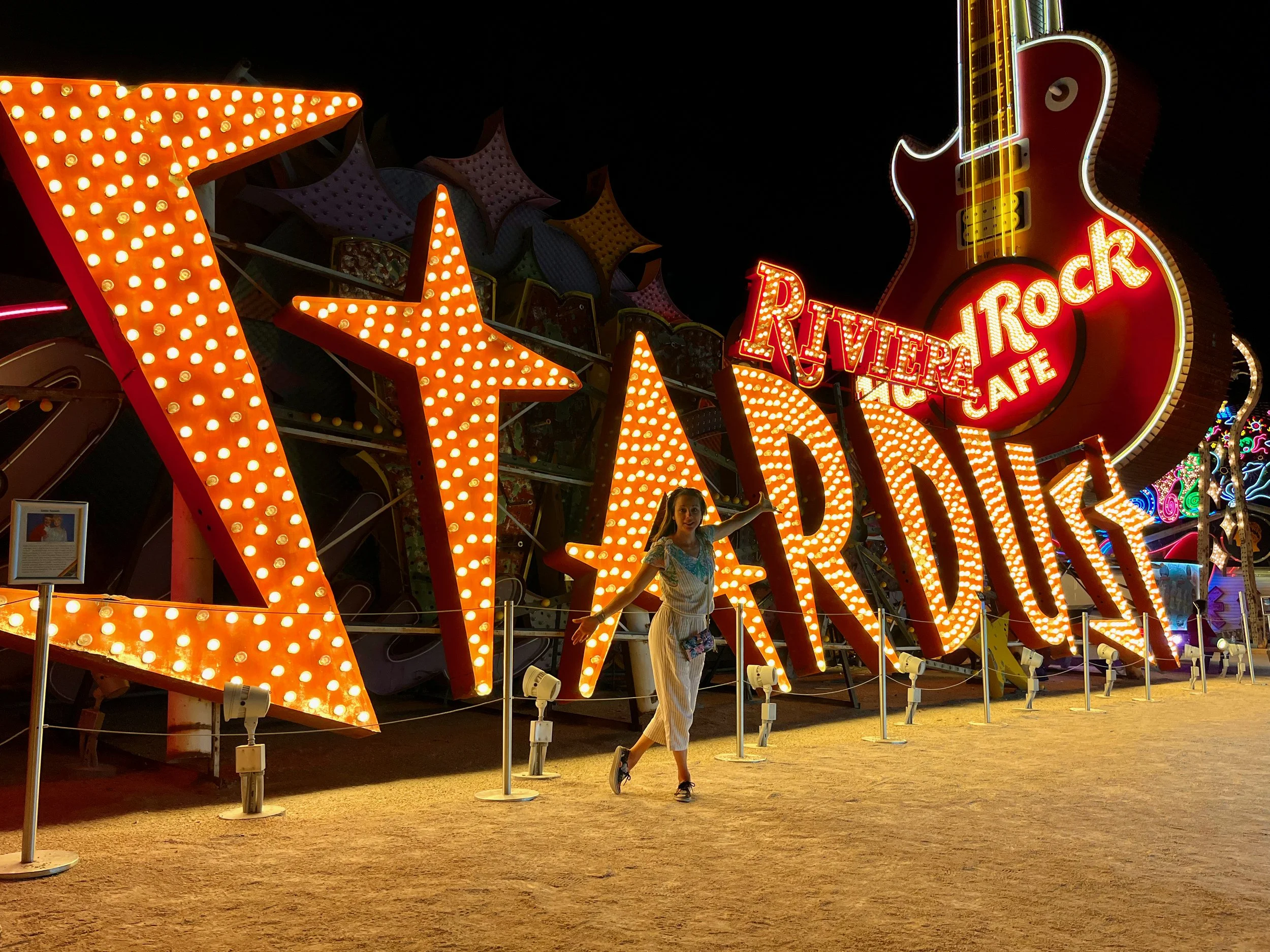 Roberta in a light-colored dress jumps in front of a brightly lit sign at a fair, with large illuminated stars and the words "VINTAGE" and "RIVER ROCK CAFE" in the background. Neon Museum, Las Vegas Nevada