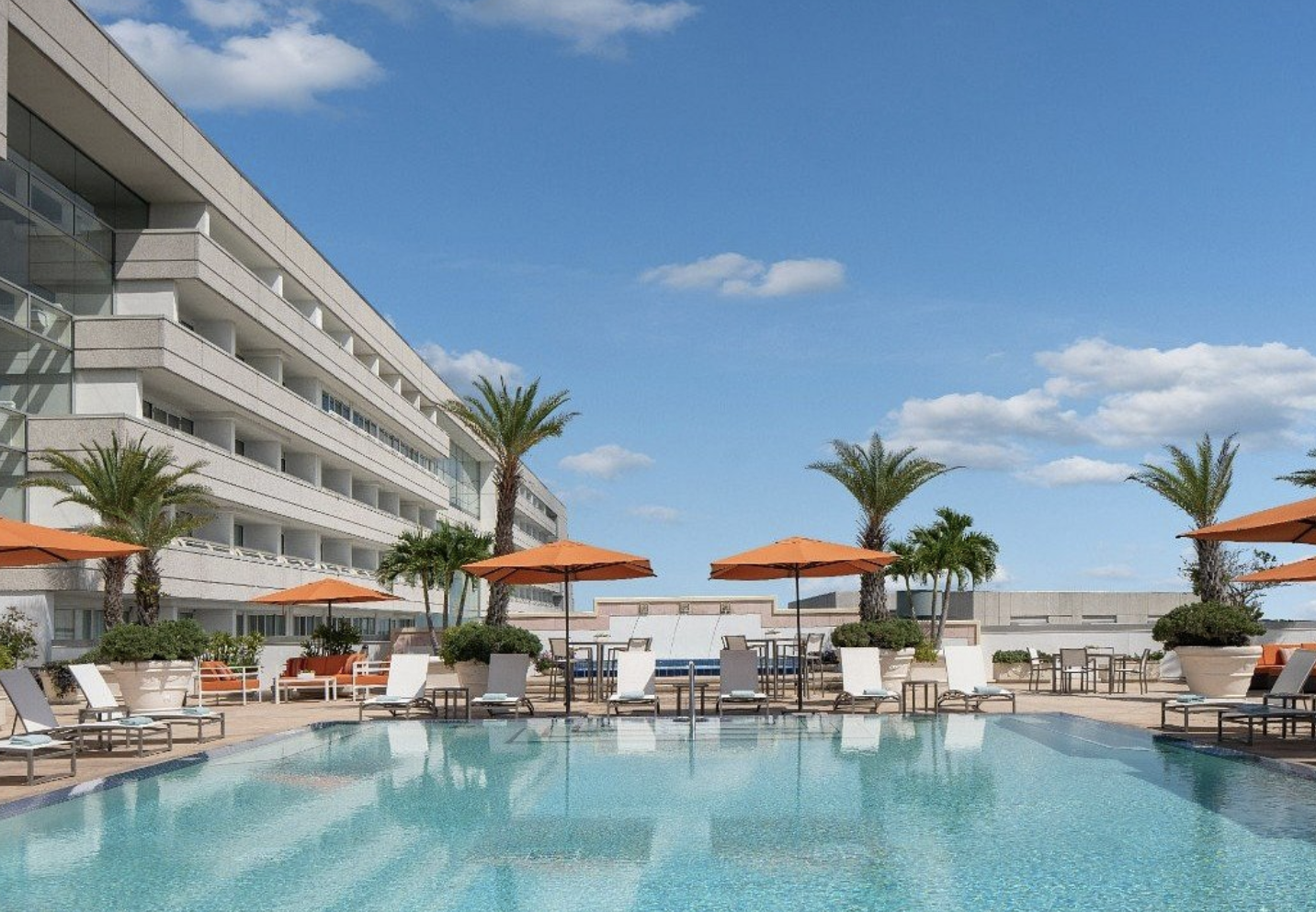 A swimming pool area at a hotel with lounge chairs and orange umbrellas, surrounded by palm trees and a multi-story hotel building under a blue sky with some clouds.