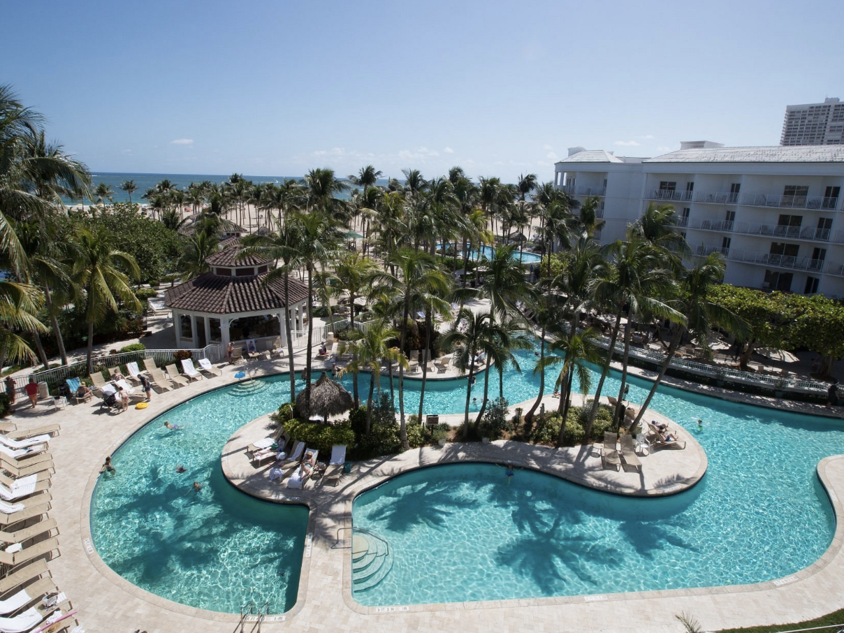 Resort swimming pool area with palm trees, lounge chairs, and a gazebo, overlooking the ocean on a sunny day.