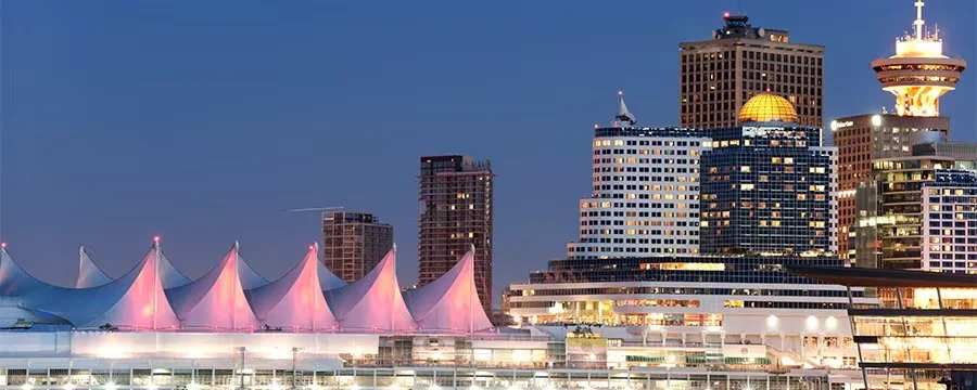 Nighttime view of a city skyline featuring modern high-rise buildings and a waterfront with a stadium that has a white, tent-like roof illuminated with pink lights.