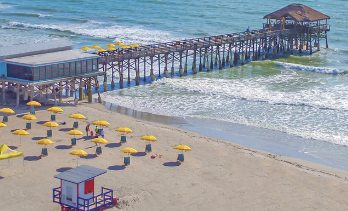 Beach scene with yellow umbrellas, a pier extending into the ocean, and a sandy shore with a lifeguard tower.