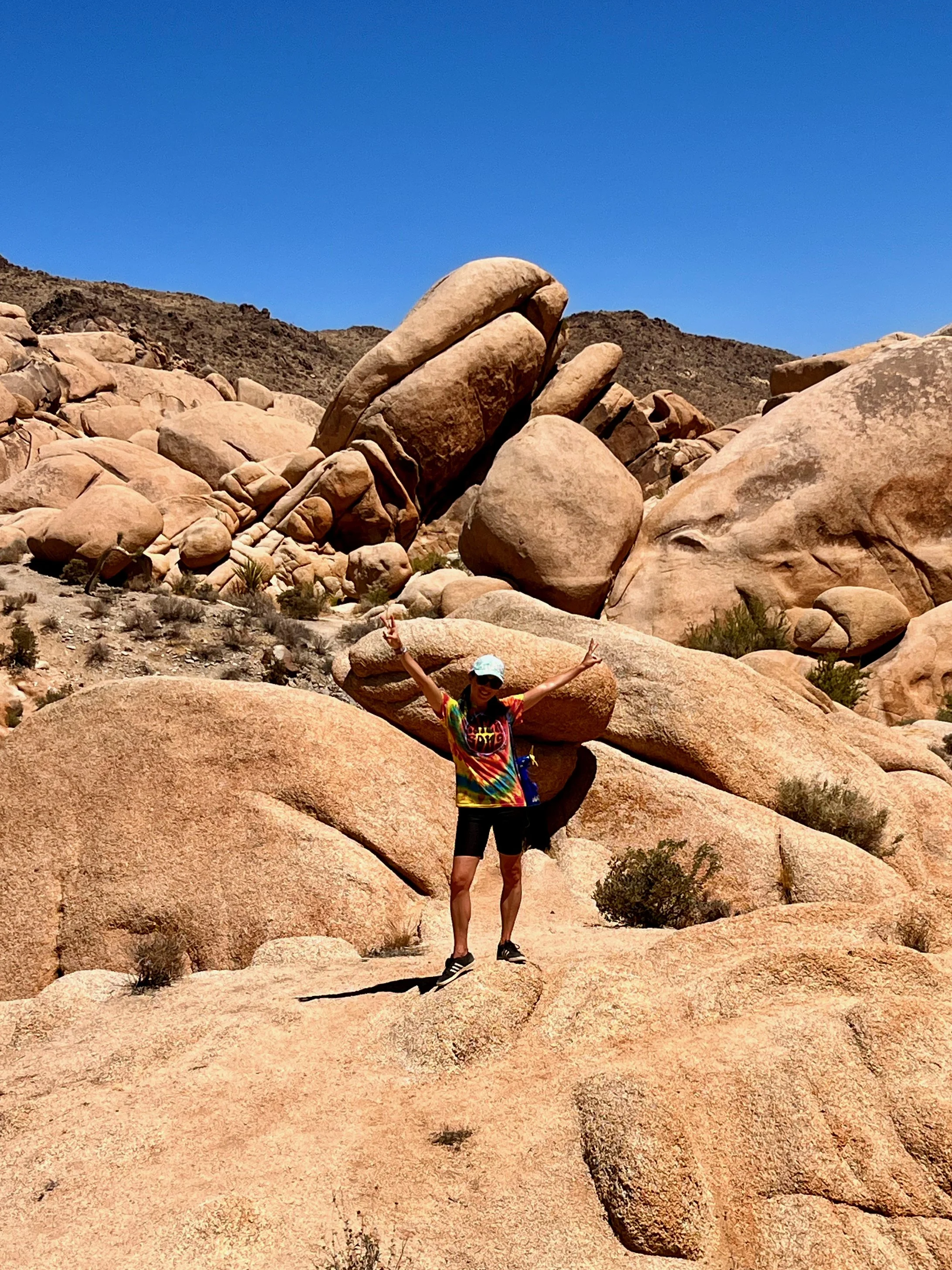 Roberta wearing an Earth Song tie-dye shirt, shorts, and a hat standing on a large desert rock formation with arms raised, surrounded by rocky mountains under a clear blue sky. Joshua Tree National Park