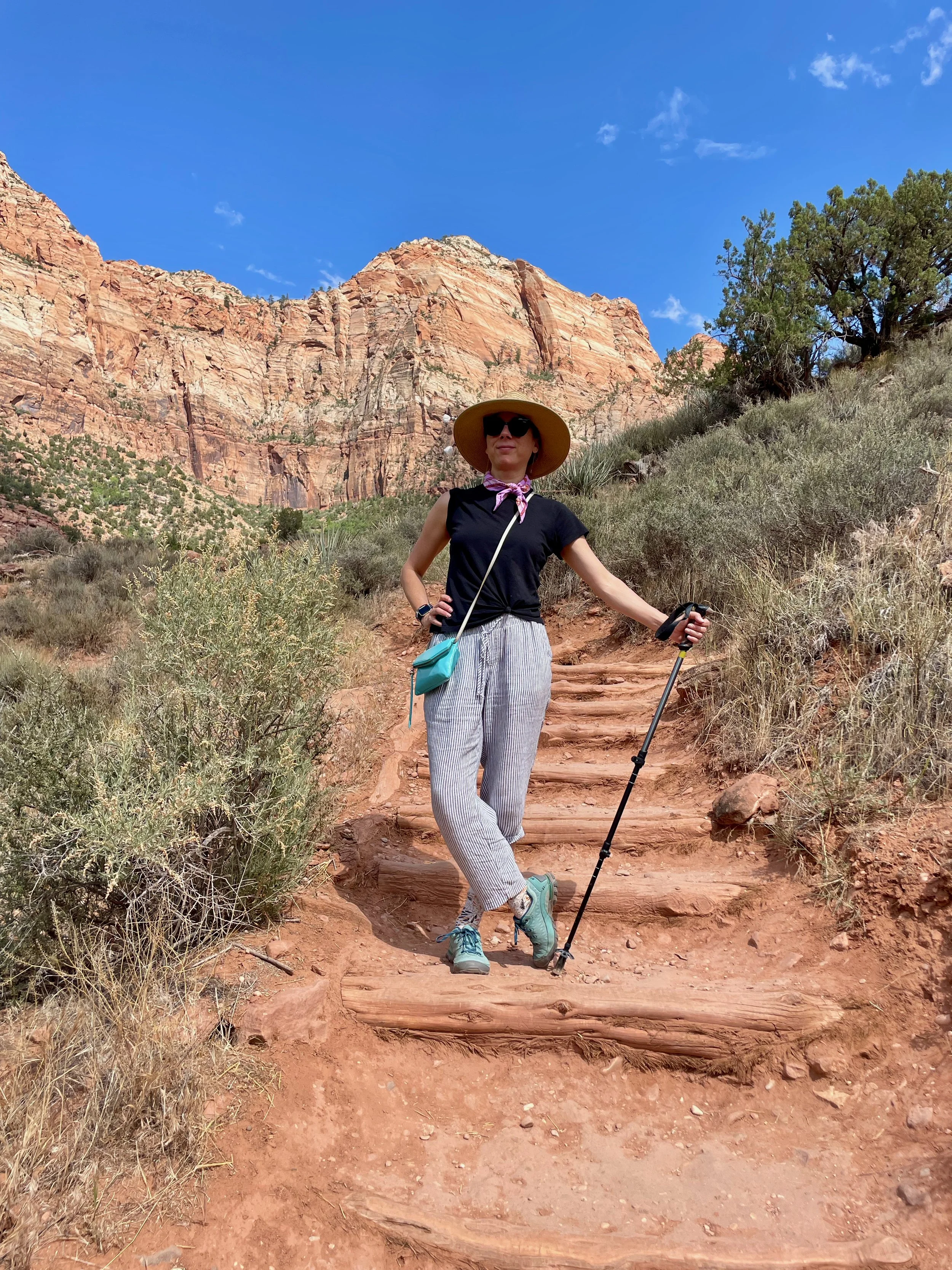 Roberta hiking on a dirt trail with rocky, reddish-brown mountains and blue sky in the background, wearing a wide-brimmed hat, sunglasses, a black t-shirt, striped pants, and hiking boots, holding a trekking pole. Sedona Arizona