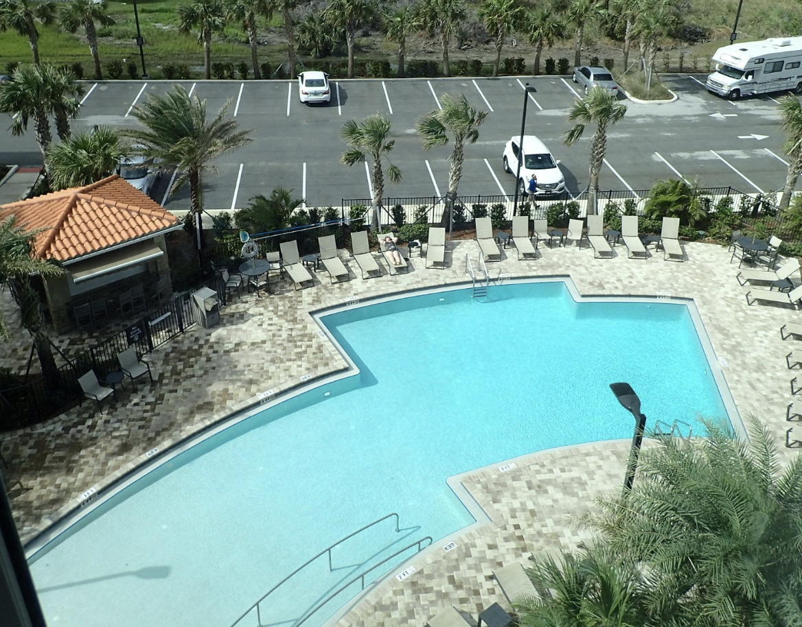 An aerial view of a hotel swimming pool surrounded by lounge chairs, palm trees, and a parking lot with cars and an RV camper.