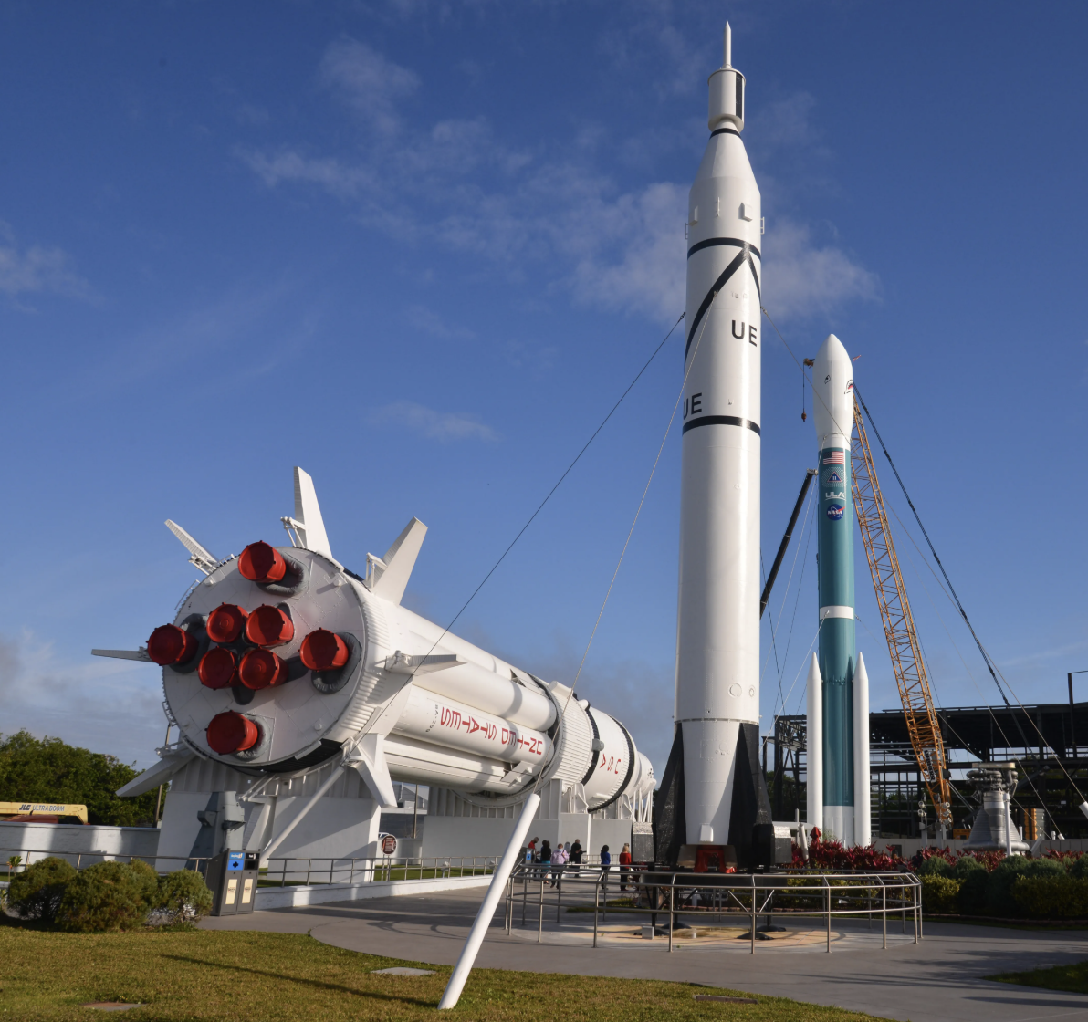 Multiple rockets displayed outdoors, with one rocket on a launch pad, a large rocket standing upright, and another smaller rocket with a crane beside it.