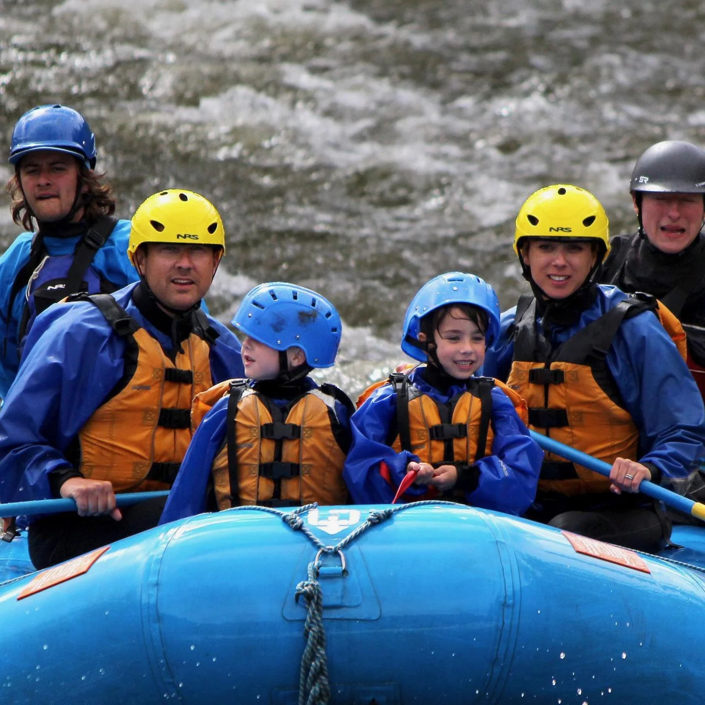 Roberta and her family, are on a rafting trip in a river, wearing helmets and life jackets, with water splashing in the background. Keystone, Colorado