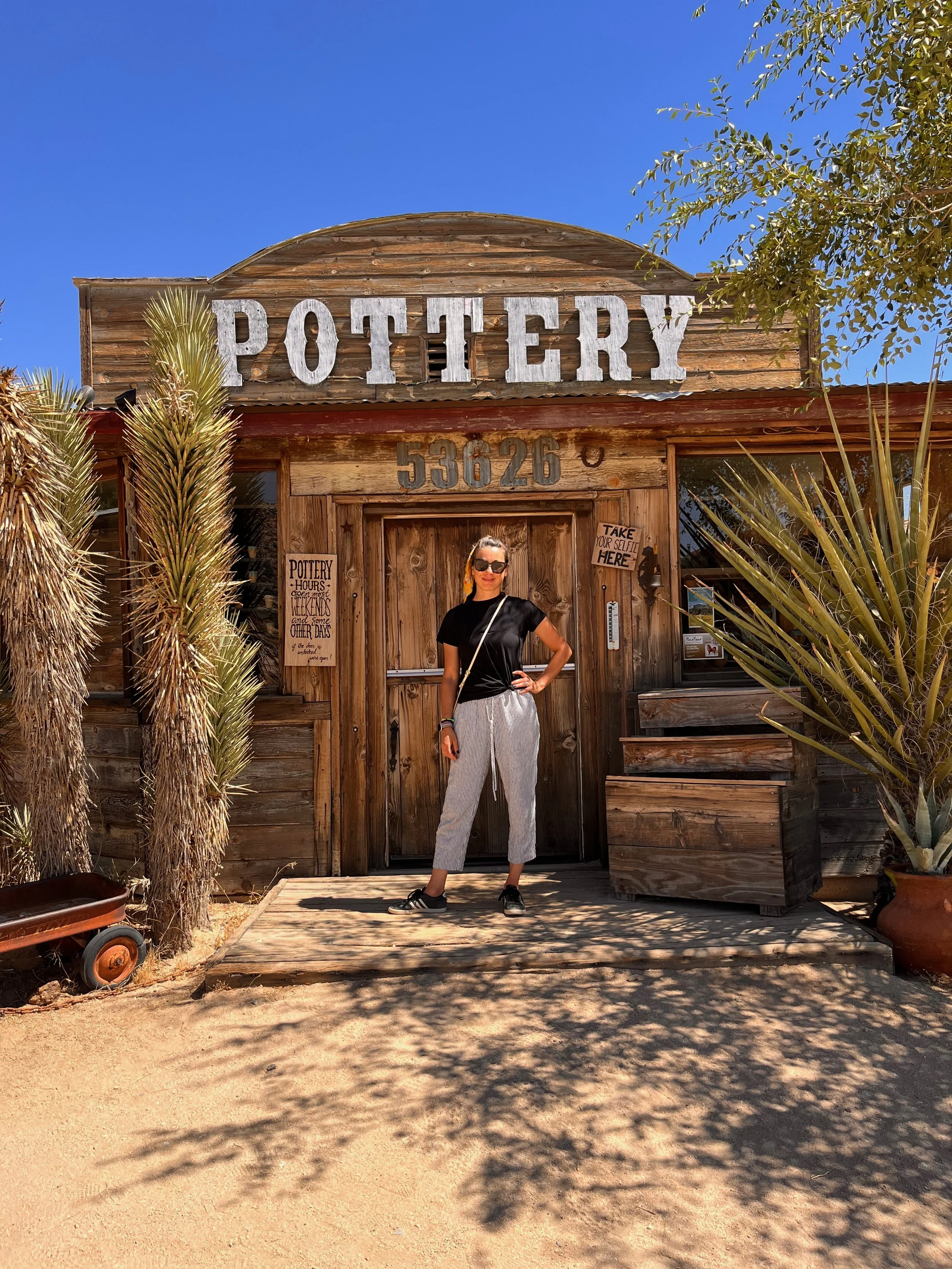Roberta standing in front of a rustic pottery shop with a wooden exterior, large 'POTTERY' sign, and greenery around. She is wearing sunglasses, a black t-shirt, striped pants, and sneakers, with her hand on her hip. Pioneer Town, California