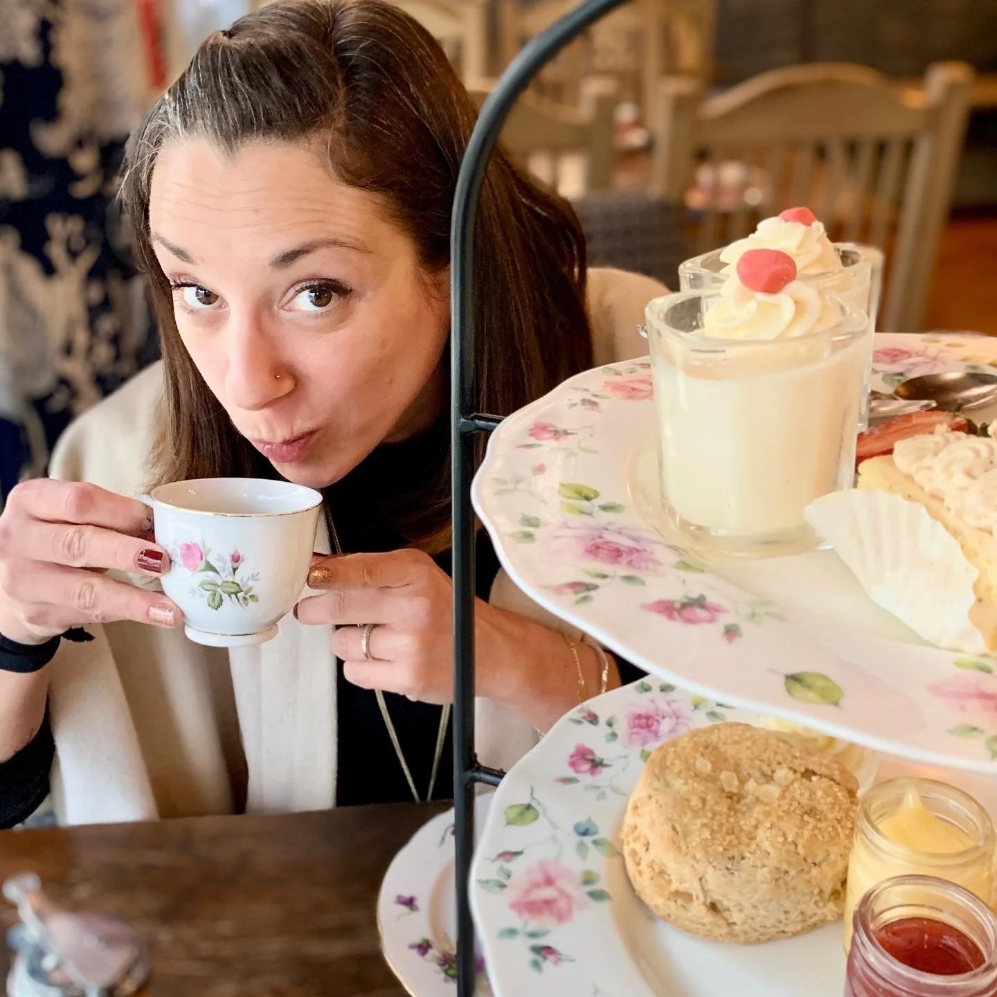 Roberta holding a teacup decorated with pink and green floral pattern, looking at the camera, with a three-tiered tray of desserts and scones in front of her in a cozy café setting.