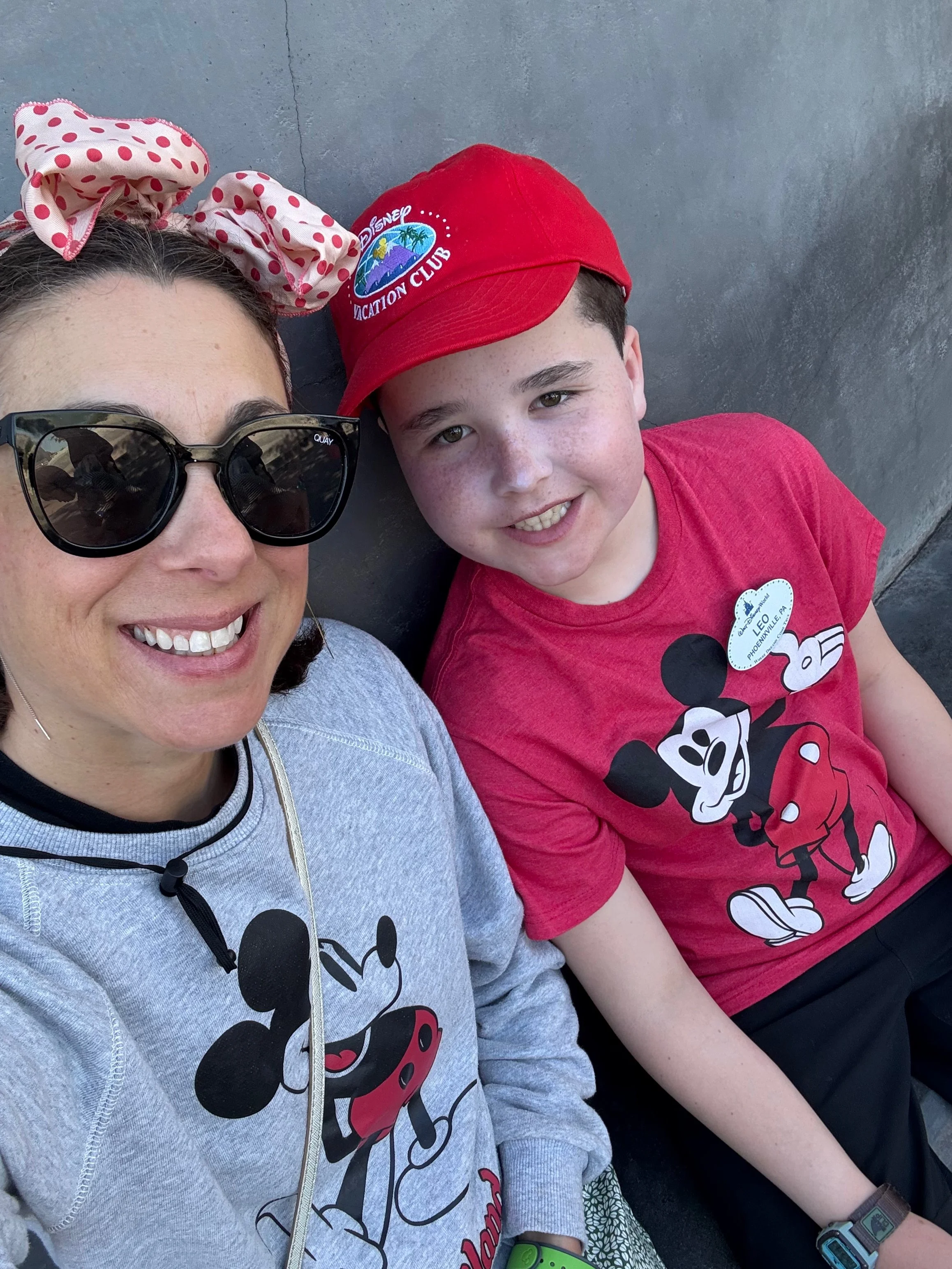 Roberta smiling woman wearing sunglasses and a gray Mickey Mouse sweatshirt sitting next to a smiling boy in a red Mickey Mouse shirt and red Disney Vacation Club hat, both outdoors against a concrete wall.