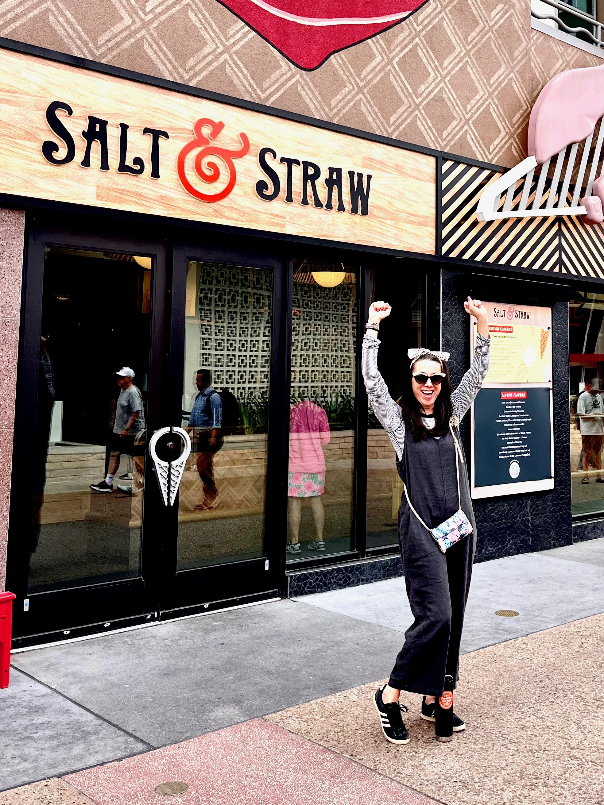 Roberta with sunglasses, a hair bow, and black overalls standing outside a Salty & Straw restaurant with arms raised in celebration. Best Ice Cream in Disney