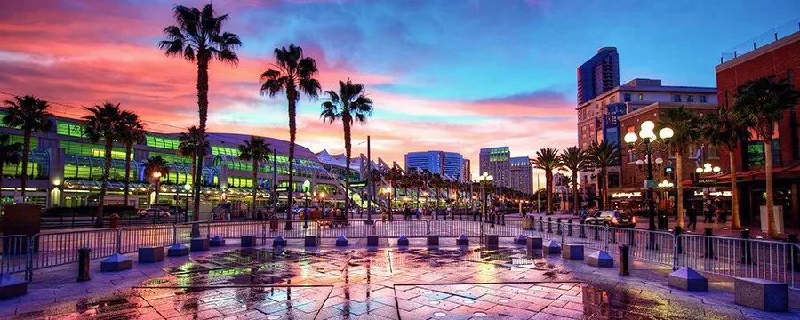 Sunset over a lively city street with palm trees, colorful sky, and illuminated buildings reflected on wet pavement
