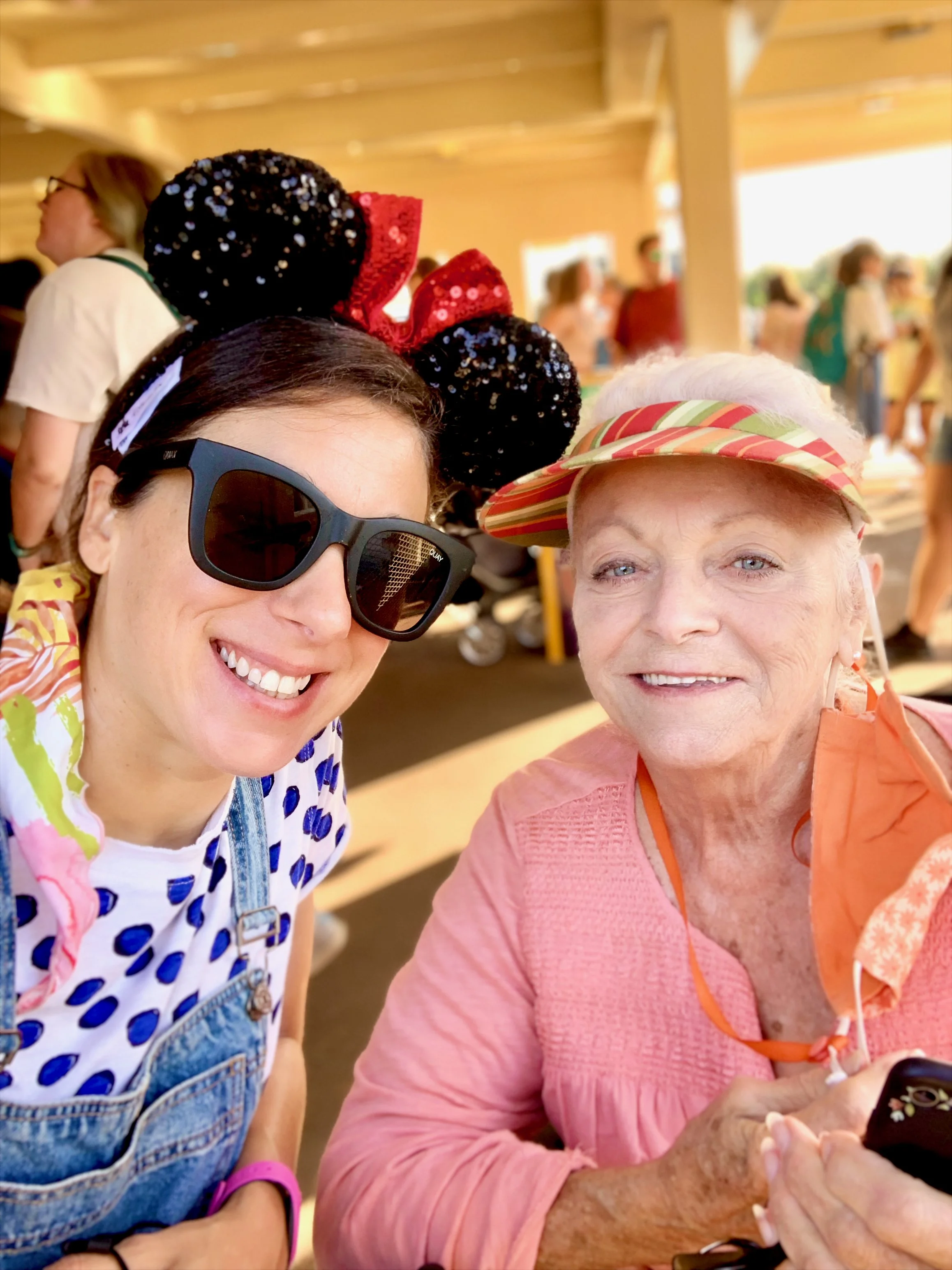 Roberta and her Mom smiling for a selfie, one wearing black sunglasses and Mickey Mouse ears headband, the other wearing a striped visor and orange shirt, at an outdoor event under a wooden roof with people in the background.
