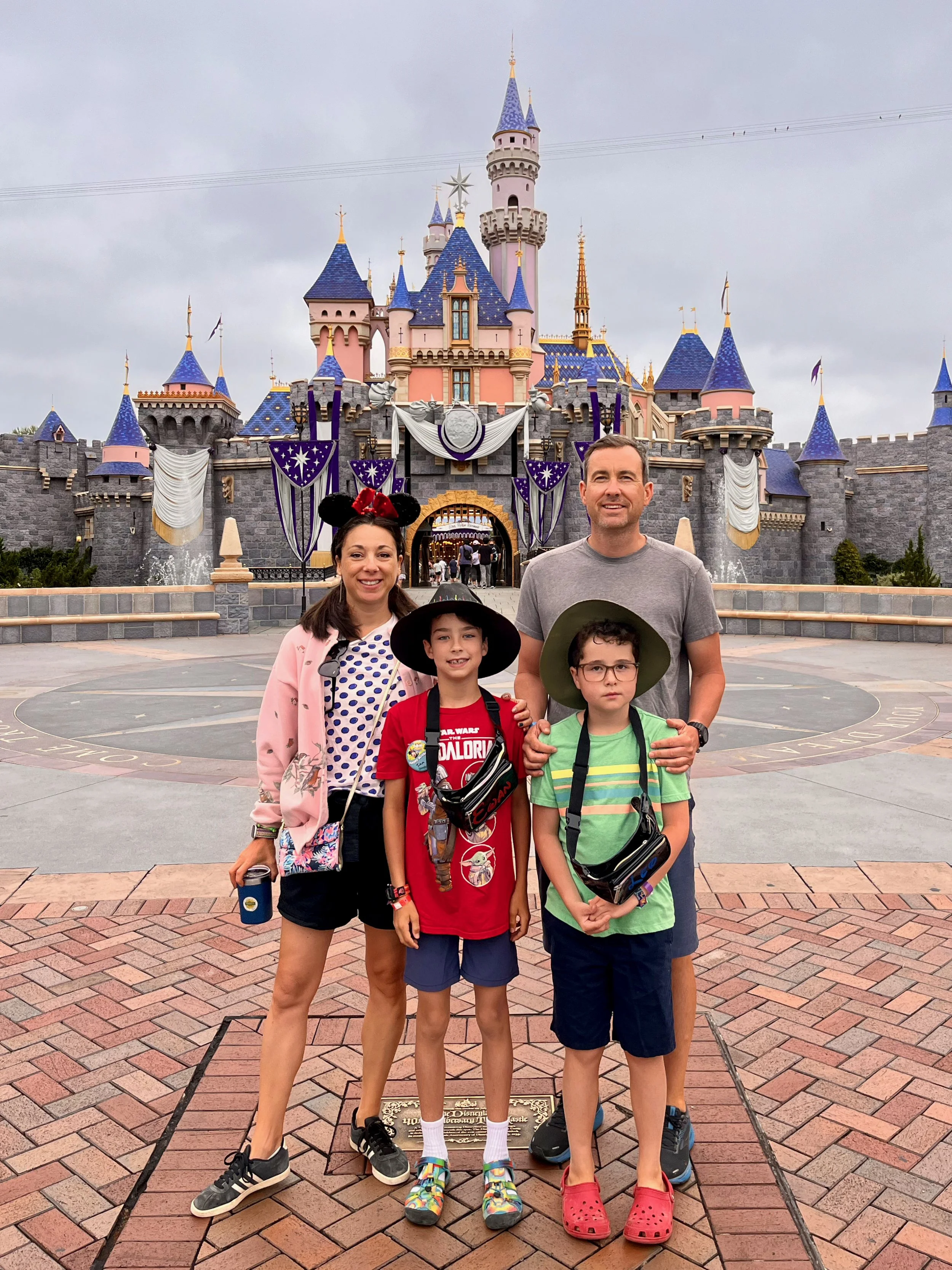 Roberta and her family posing in front of Sleeping Beauty Castle at Disneyland, with cloudy sky and park entrance in the background.