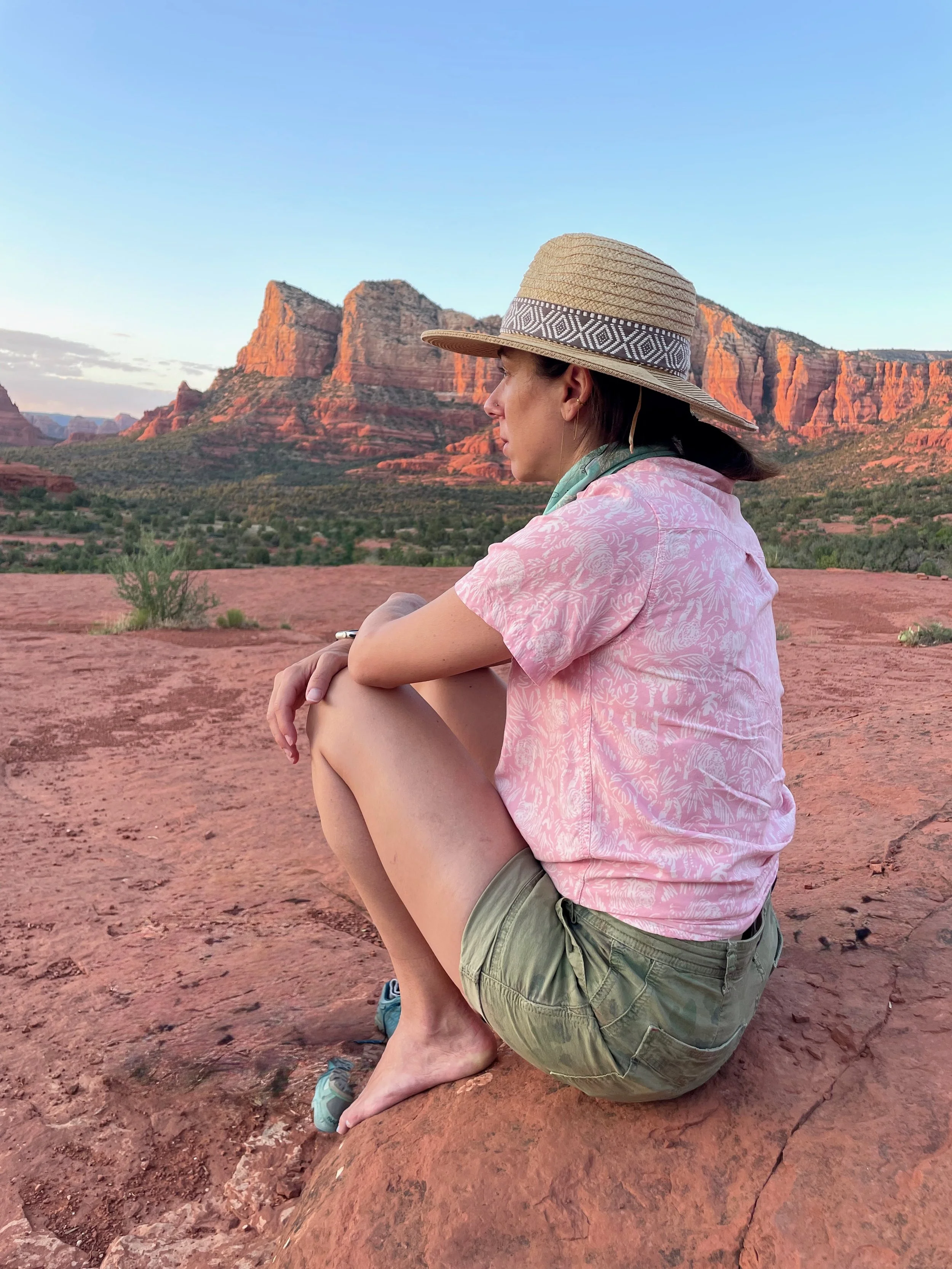 Roberta wearing a large sunhat, pink patterned shirt, and beige shorts sitting on a reddish rock with her knees drawn up, overlooking the red rock formations and desert landscape of Sedona, Arizona during sunset.