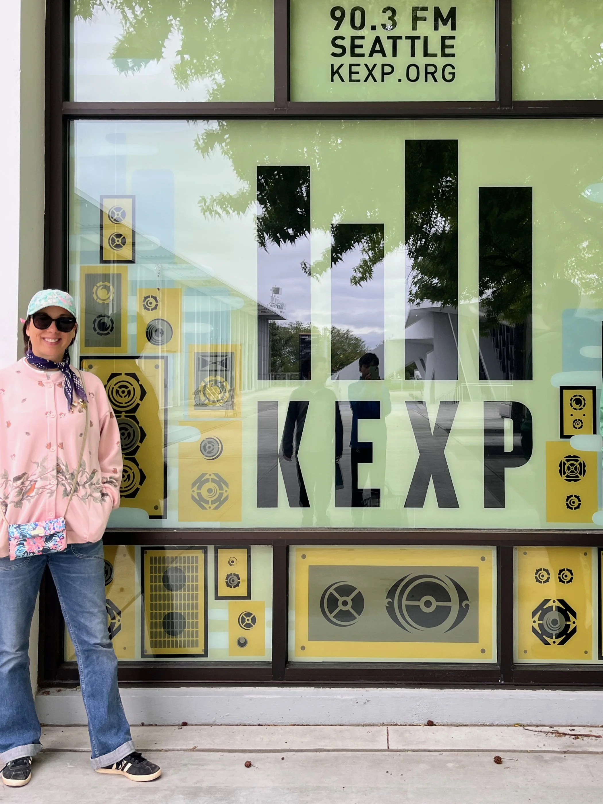 Roberta standing in front of a large window with graphics and text promoting KEXP radio station, with trees and cloudy sky reflected in the window. Seattle Washington