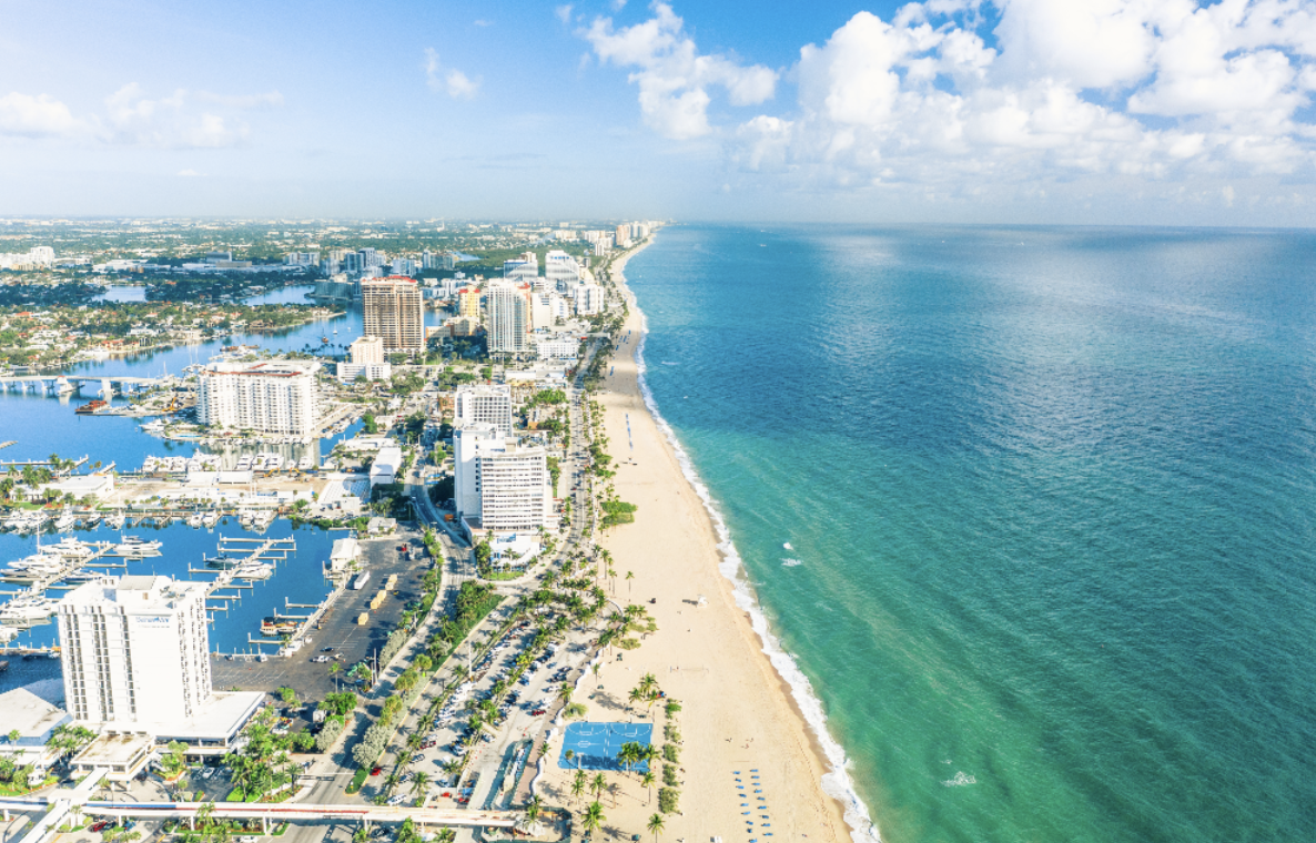 Aerial view of a coastal city with a sandy beach, high-rise buildings, boats in a marina, and turquoise ocean waters under a partly cloudy sky.