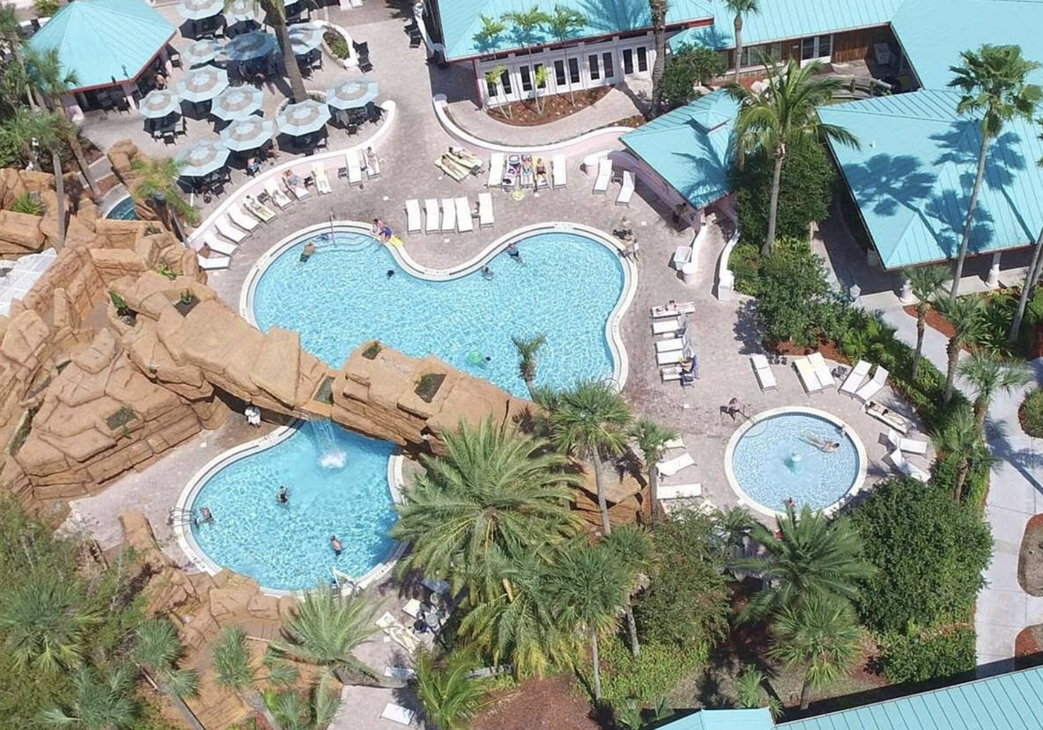 An aerial view of a resort swimming pool area with multiple pools, surrounded by lounge chairs, umbrellas, palm trees, and shaded structures.