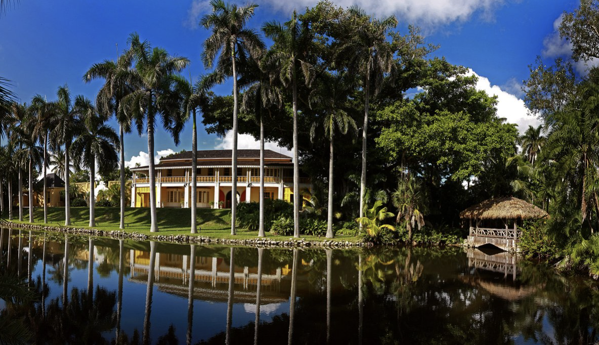 A tropical landscape with a large house, tall palm trees, lush green trees, a pond with reflections, and a thatched-roof gazebo, under a sunny blue sky with some clouds.