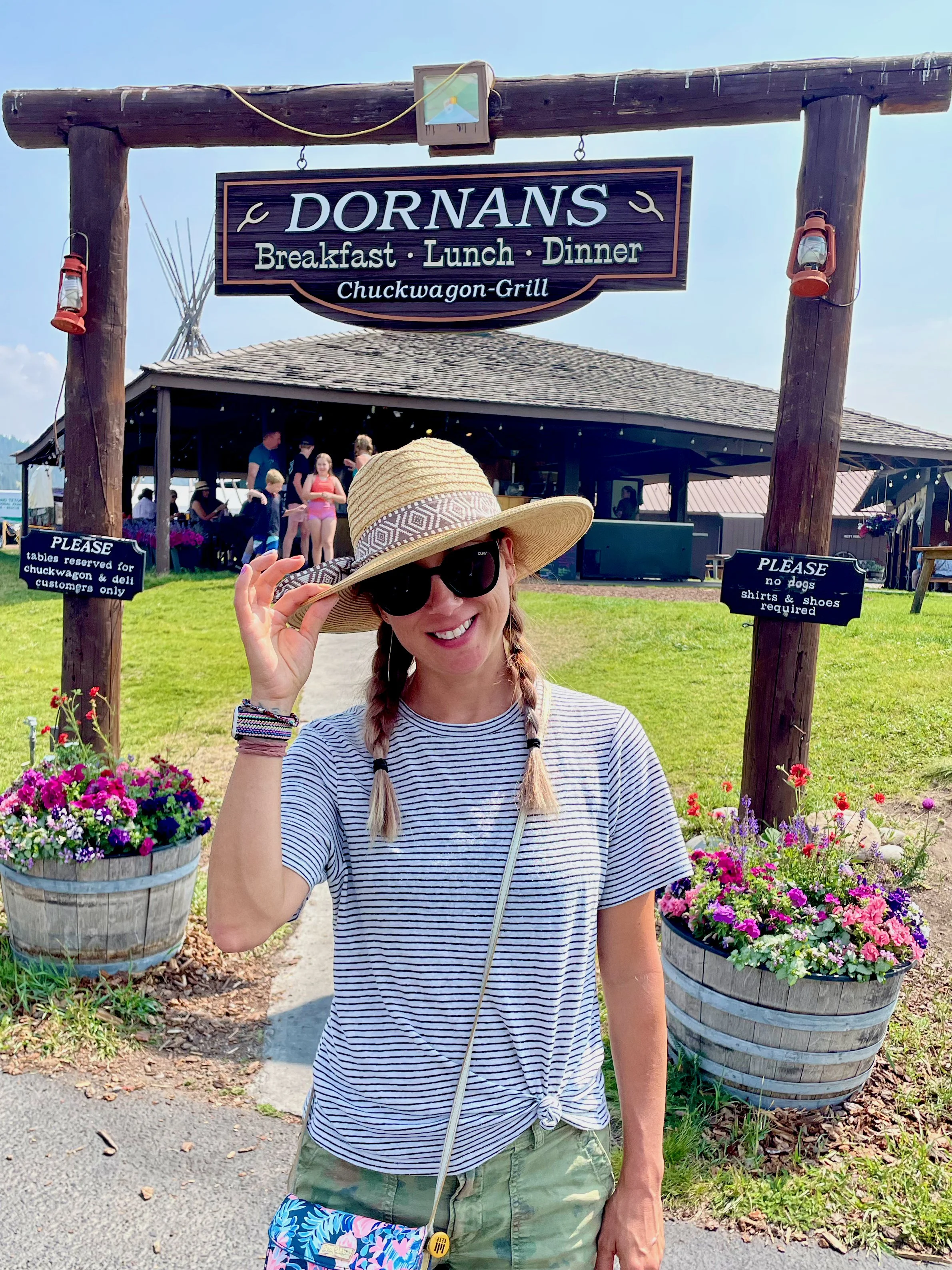 Roberta wearing a straw hat and sunglasses standing in front of a restaurant sign that reads 'Dornans Breakfast Lunch Dinner Chuckwagon-Grill.' She is smiling, wearing a striped t-shirt, and holding her hat with one hand.