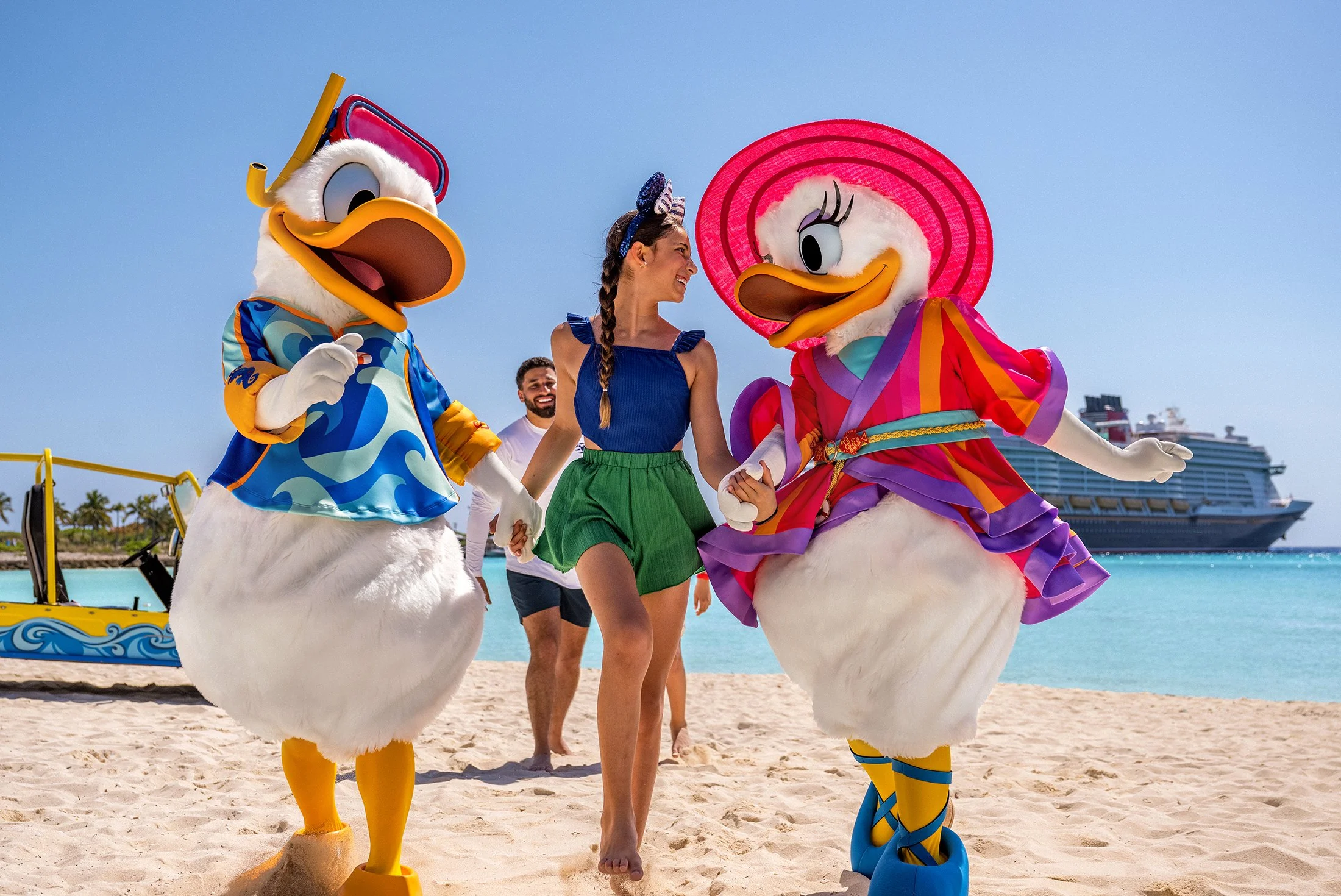 A woman walking on the beach holding hands with two people dressed as Donald and Daisy Duck characters. The woman is smiling and wearing a blue top and green shorts. The characters are in colorful outfits, and a cruise ship is visible in the background.