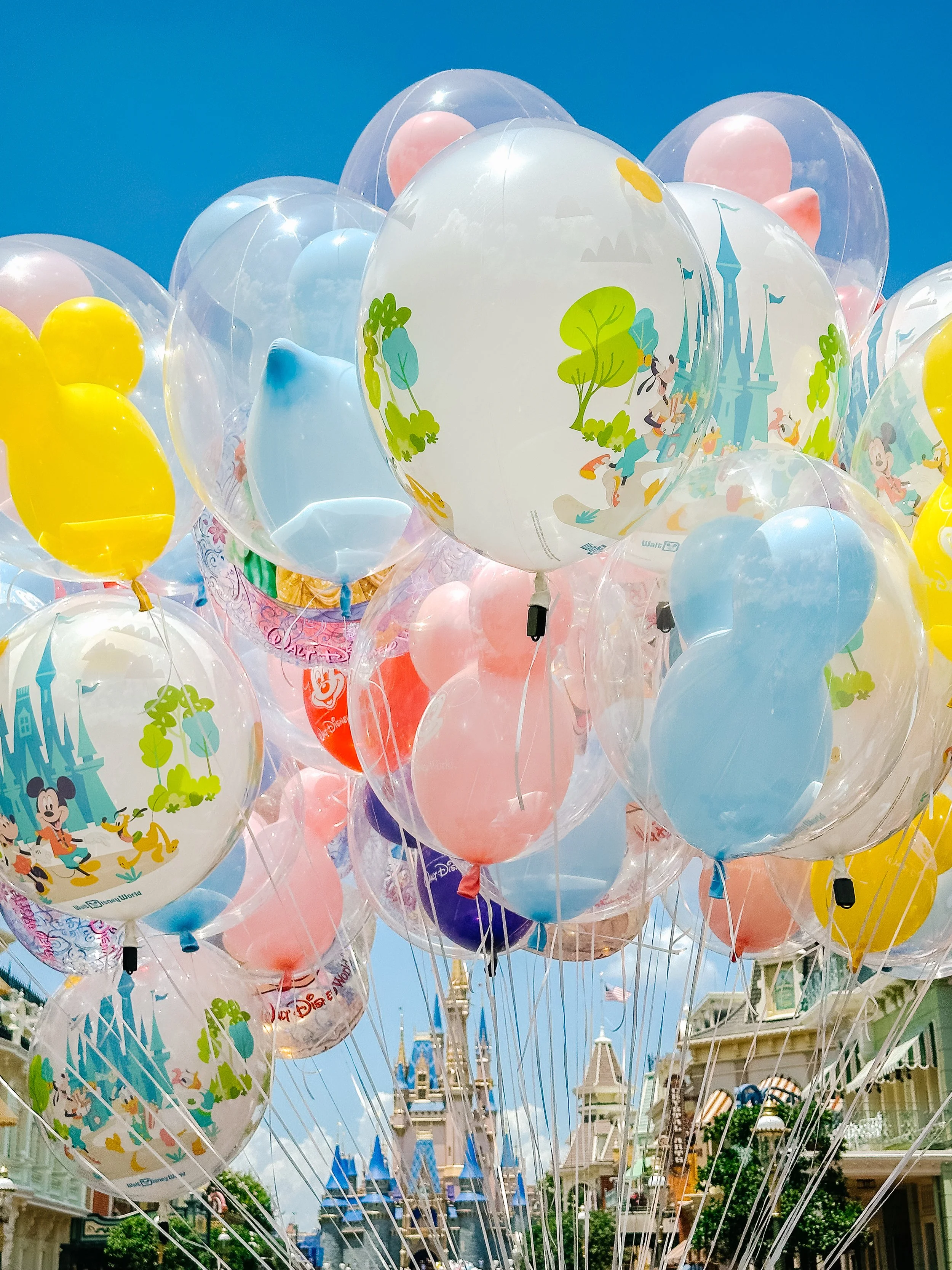 Colorful balloons featuring Disney characters and castles, with a Disney theme, held in front of Disney-themed buildings under a clear blue sky.