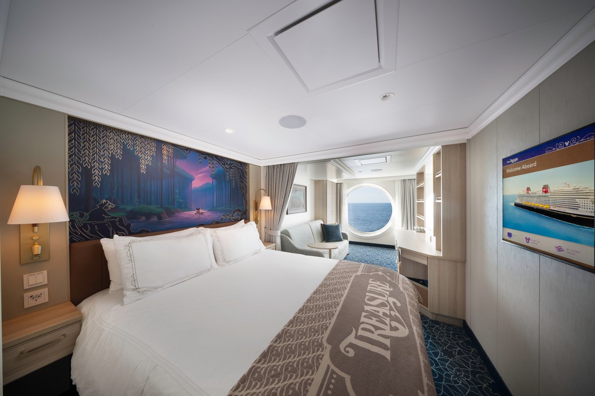Interior of a cruise ship cabin with a large bed, a window with ocean view, and a sitting area with a round window showing the sea.