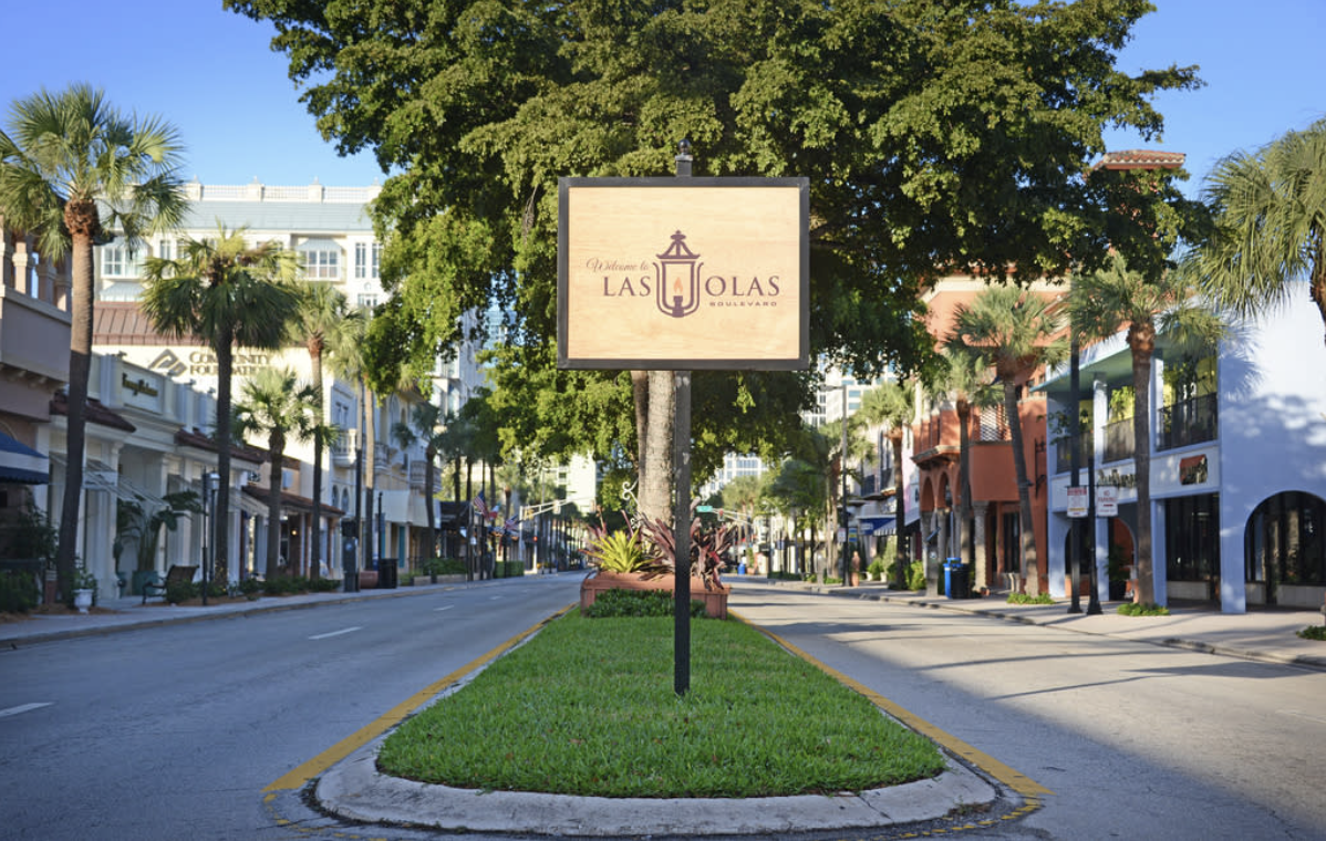 Street view of Las Olas Boulevard with a large tree in the center and shops on both sides. A welcome sign for Las Olas is in the middle of the image.