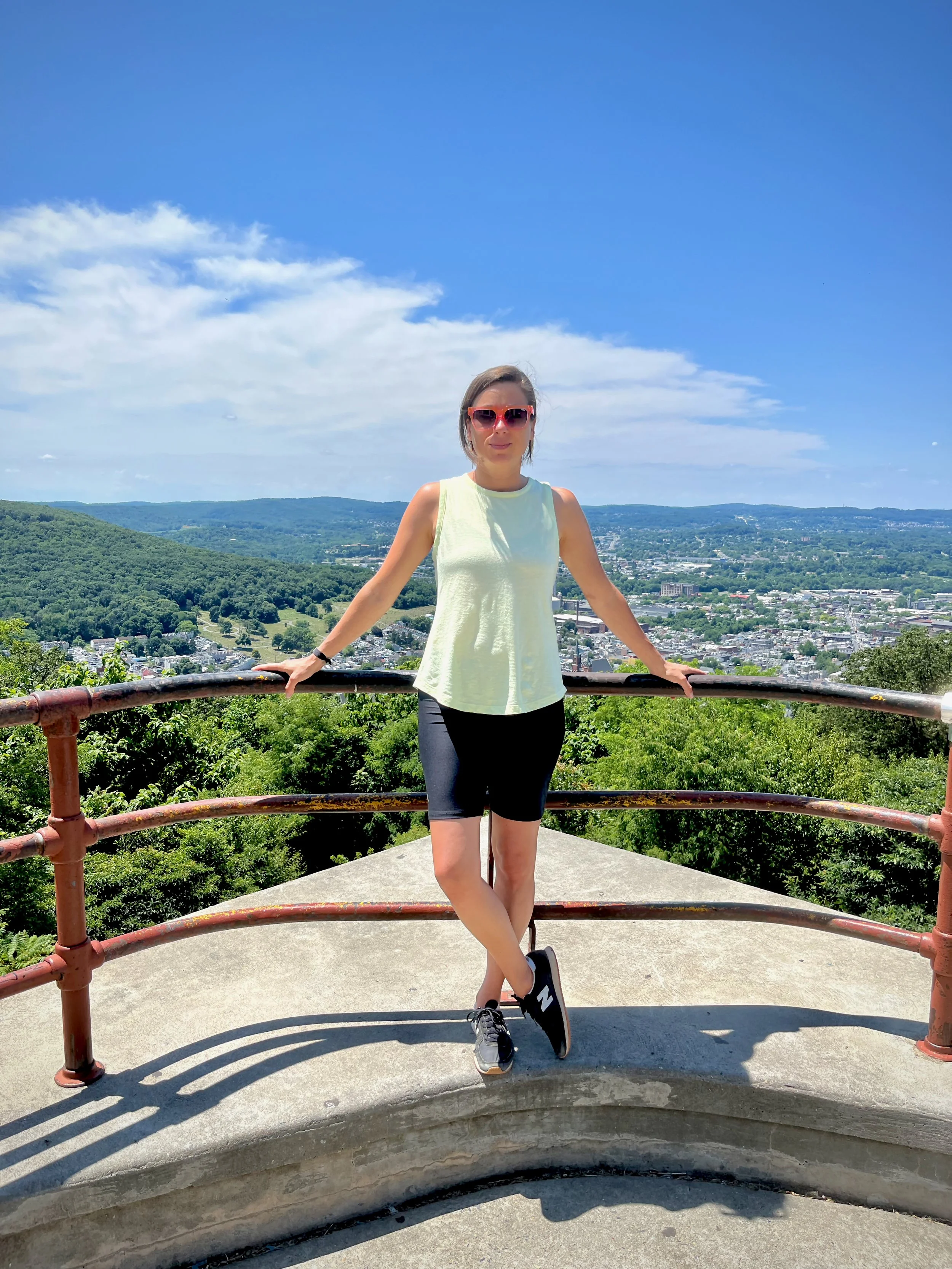 Roberta standing on a viewing platform with a scenic view of a city and green hills in the background. She is wearing sunglasses, a light-colored sleeveless top, black shorts, and sneakers. The Pagoda Reading PA