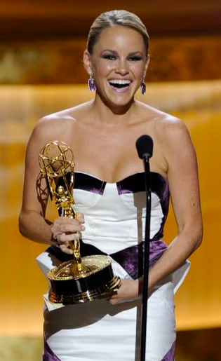 Woman in strapless dress holding an Emmy award and speaking at a microphone.