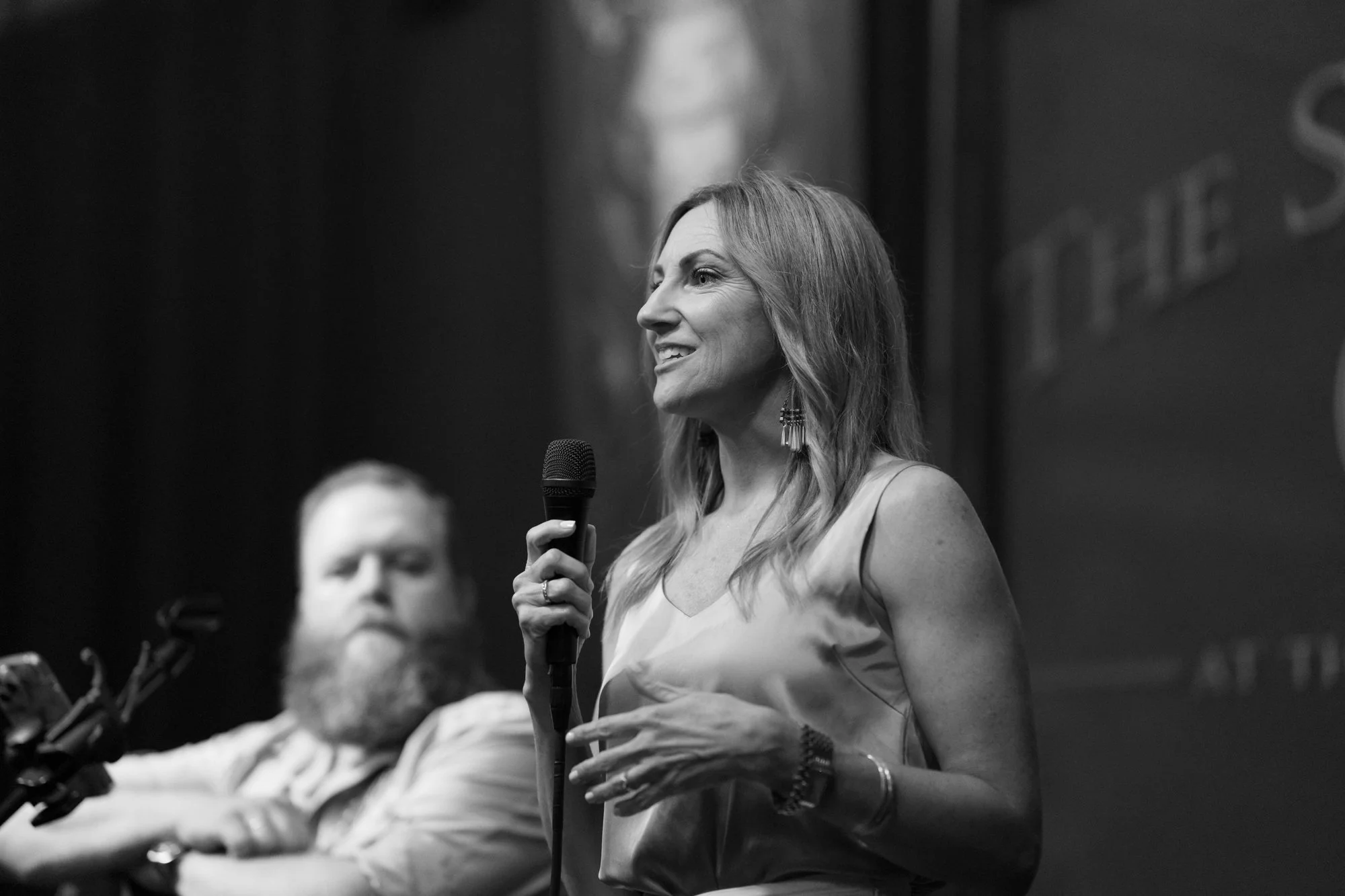 A woman speaking into a microphone on stage, with a man seated beside her, blurred in the background.