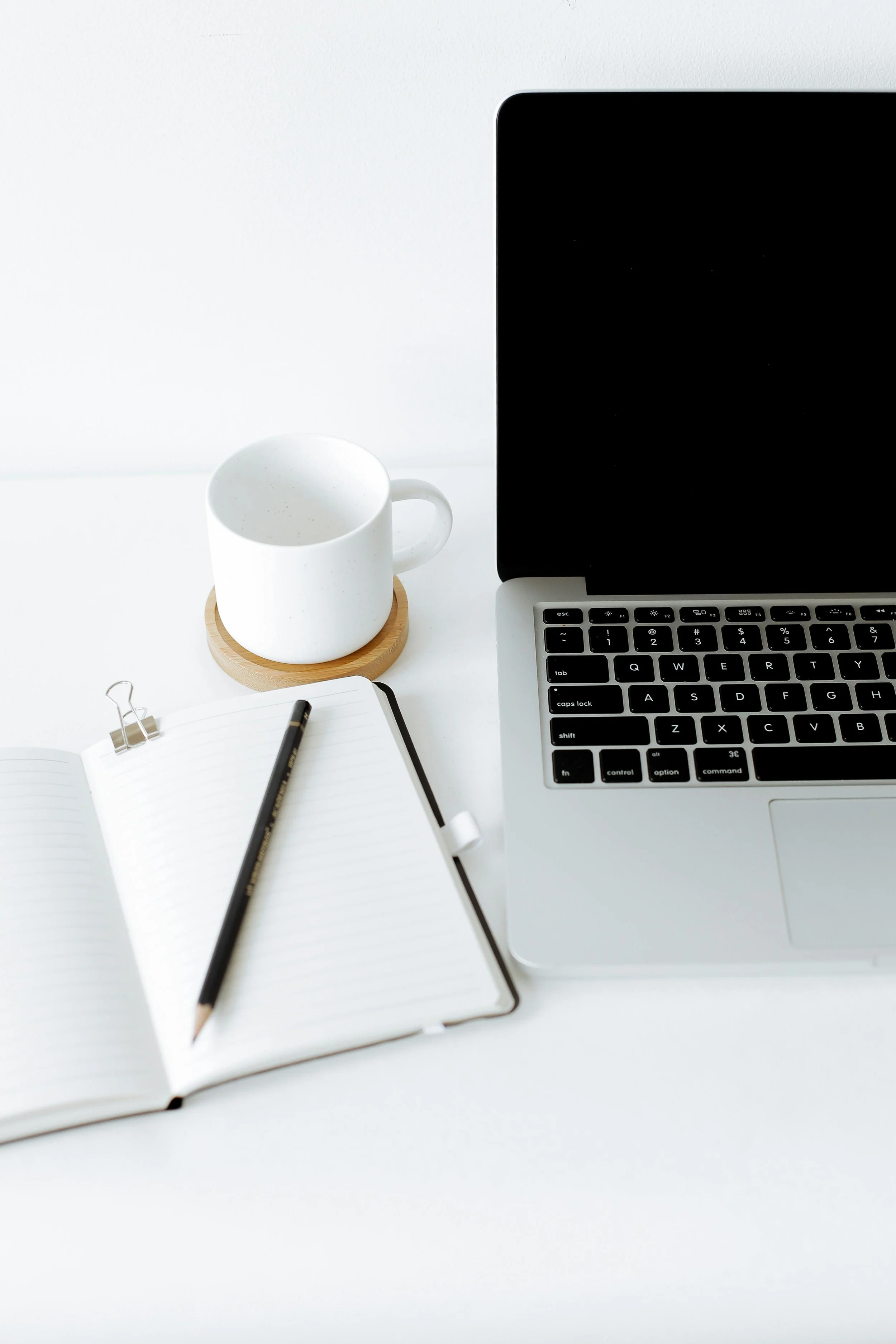 A workspace featuring a silver laptop, an open lined notebook with a black pencil, a white coffee mug on a wood coaster, and a small metal paper clip, all set on a white surface with a white background.