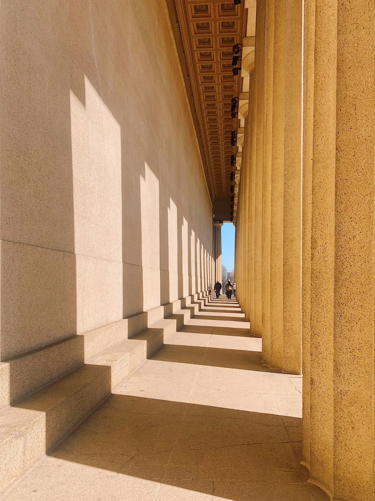 Columned walkway at the Lincoln Memorial, showing shadows cast by the columns with a few people walking in the distance.
