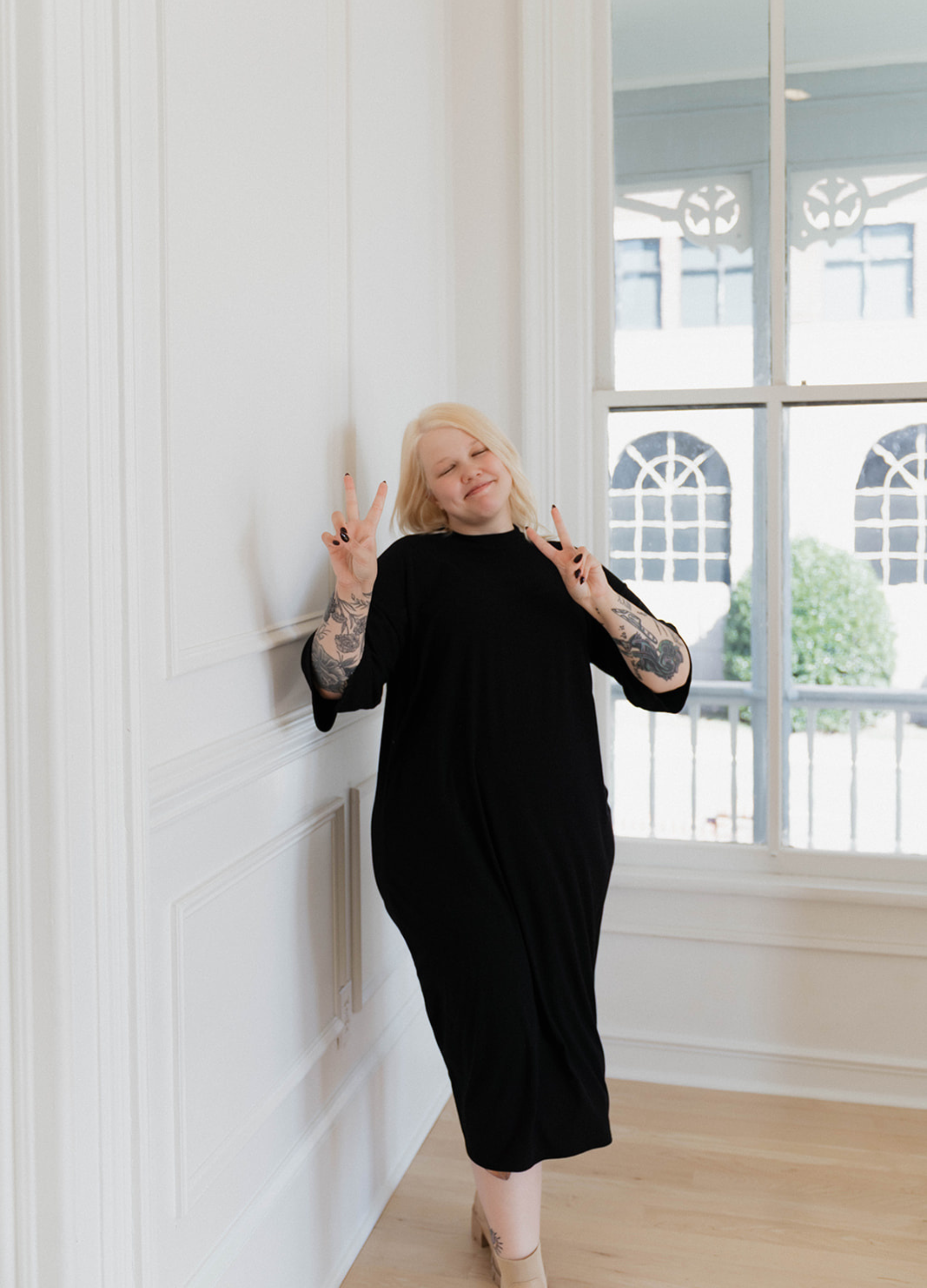 Young woman with platinum blonde hair and tattoos on her arms, standing indoors near a wall and window, making peace signs with both hands, smiling with eyes closed.