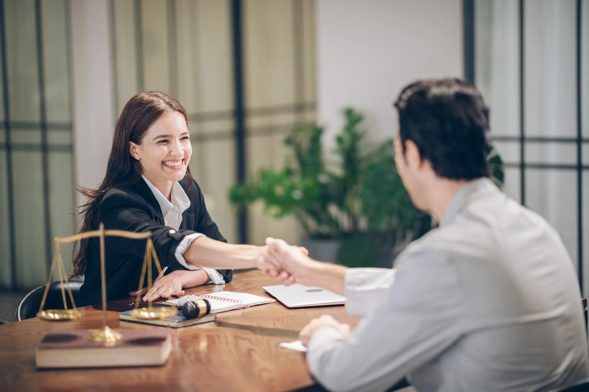 A woman in a black blazer shaking hands with a man in a white shirt in an office setting, smiling. There are legal scales, a gavel, and documents on the table.
