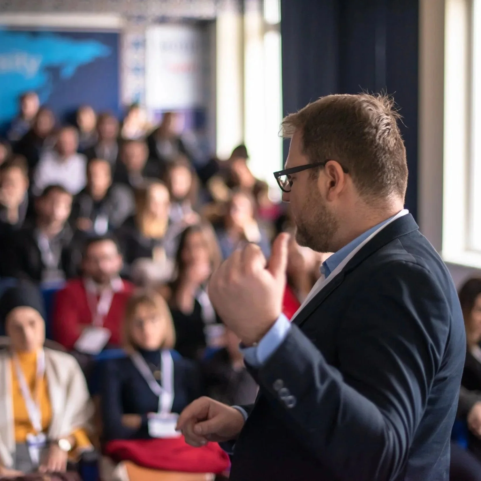 A man in a suit and glasses is giving a presentation in English in front of a seated audience in a conference room.