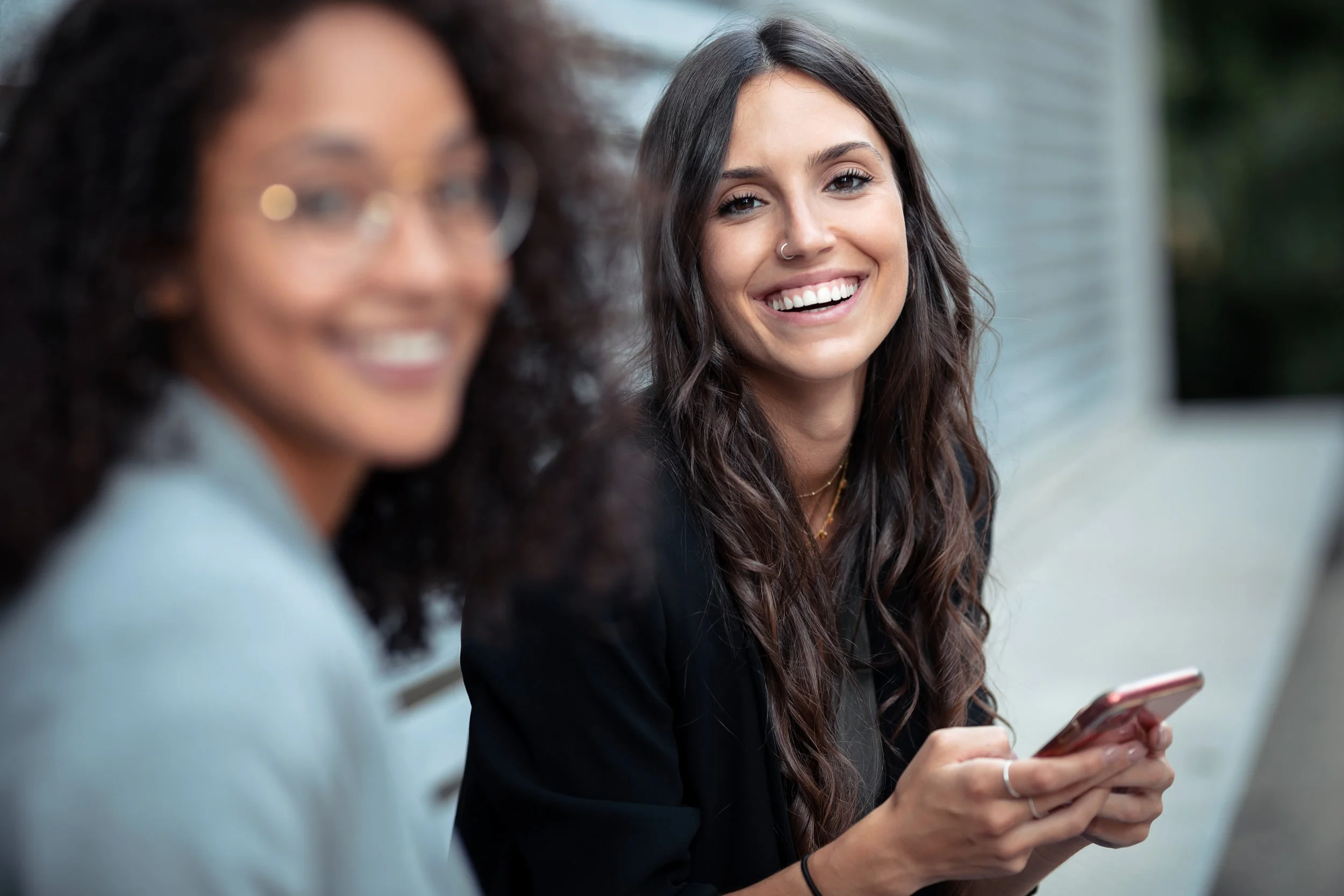 Two women sitting outdoors, one with curly hair and glasses smiling, the other with long wavy hair and a nose ring smiling while holding a smartphone.