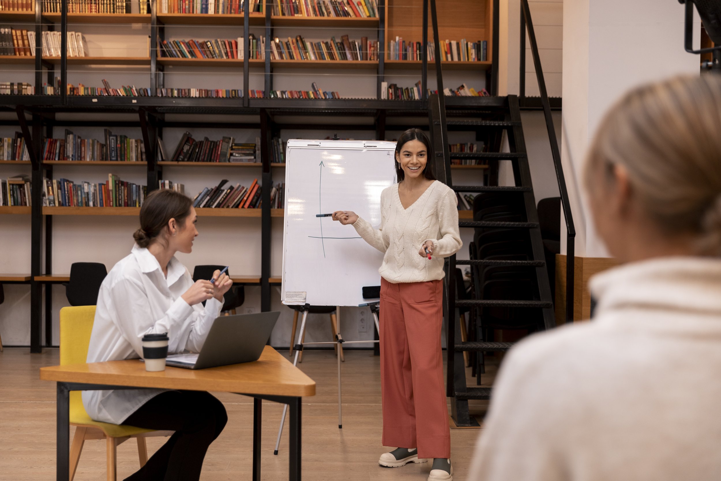 A woman standing and smiling in front of a whiteboard with a graph, holding a marker, in a modern library or classroom setting with bookshelves and stairs in the background. Two women are seated nearby, one at a desk with a laptop and coffee cup, and another in the foreground with blurred features.